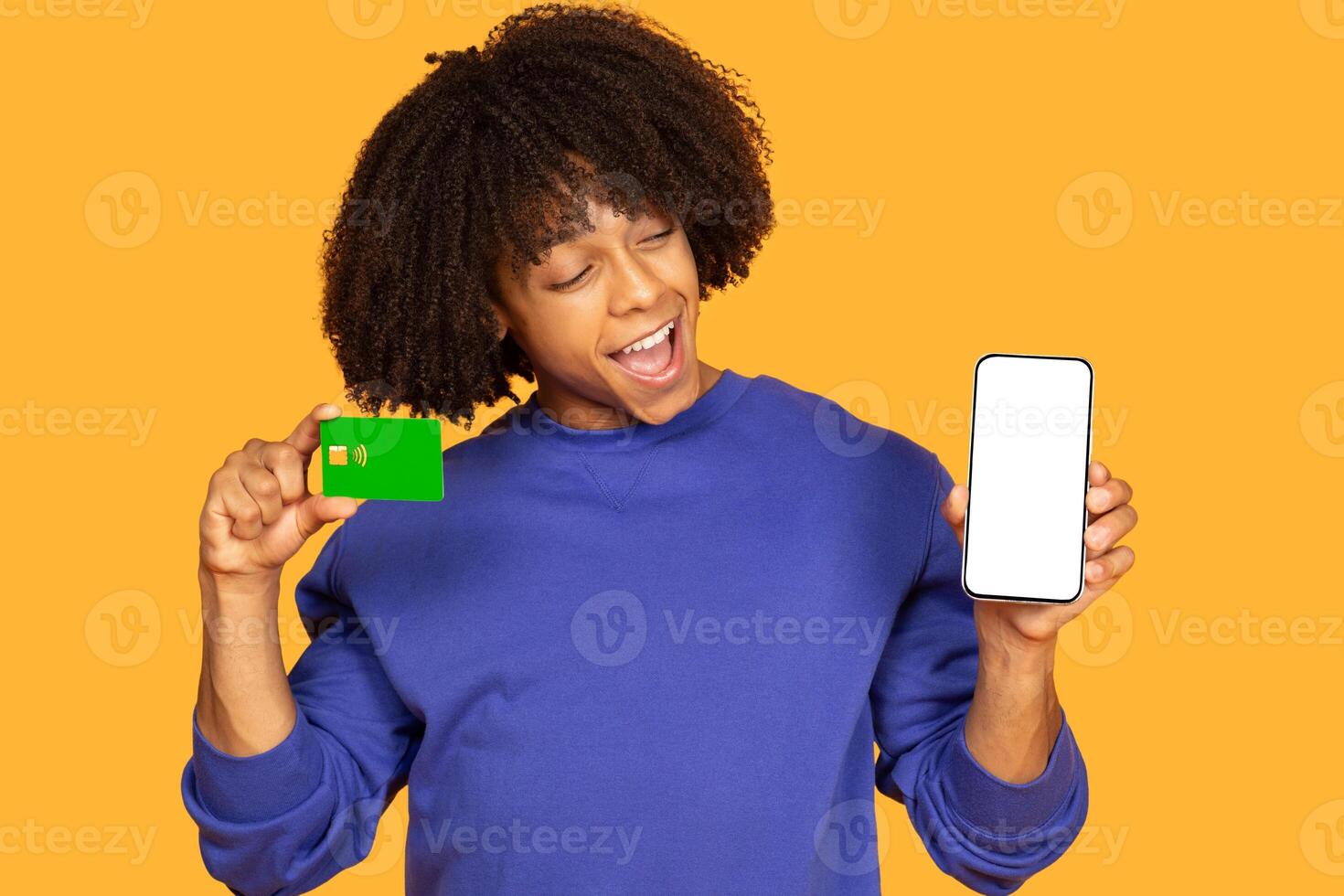 A cheerful young man poses with a mobile phone in one hand and a payment card in the other, showcasing excitement. The vibrant yellow backdrop enhances the lively atmosphere photo
