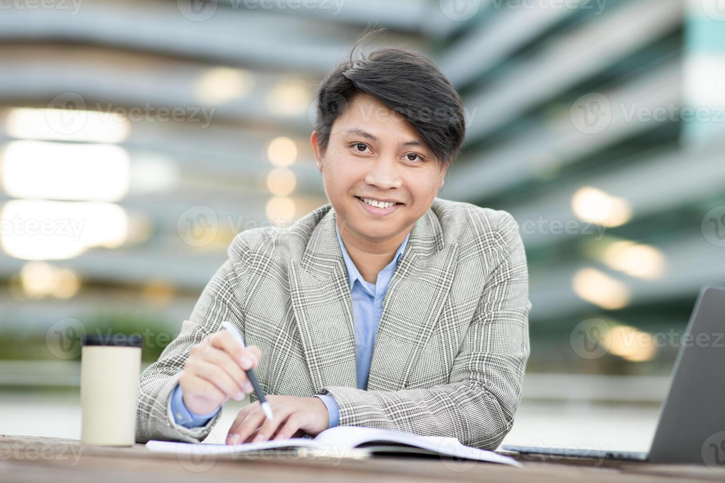 Asian man wearing a plaid jacket is sitting outside a modern building. He is writing in a notebook and looking directly at the camera with a smile. A laptop and coffee cup are on the table photo