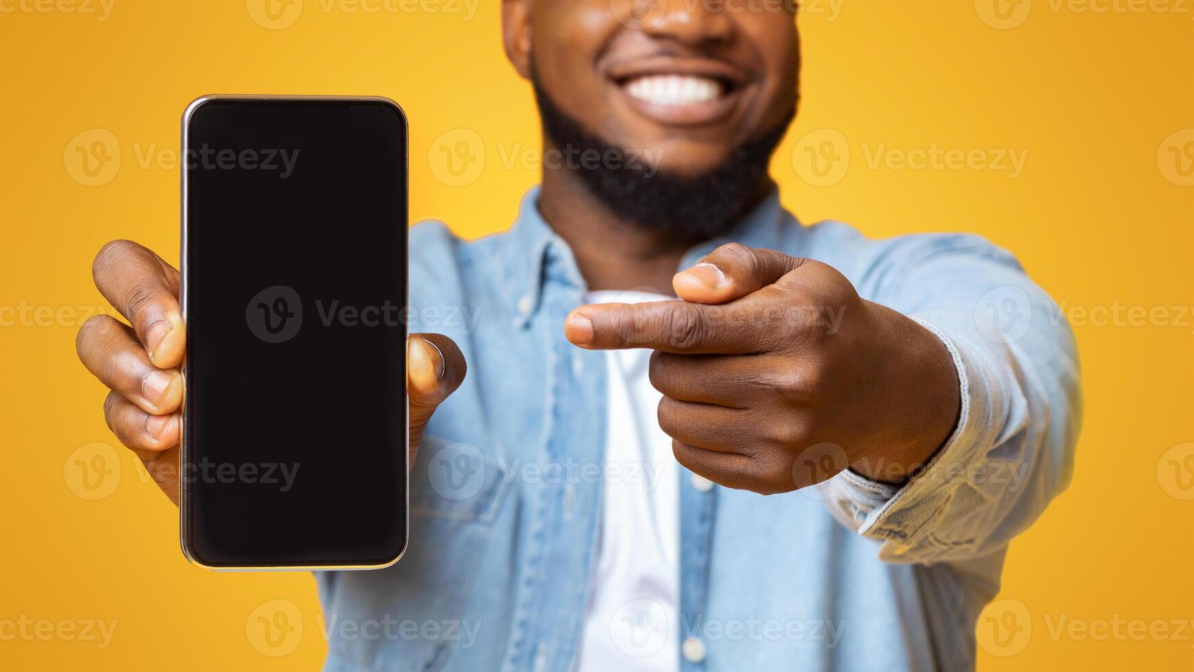 Check this app. Cheerful afro guy holding and pointing at smartphone with black screen, yellow background with free space photo