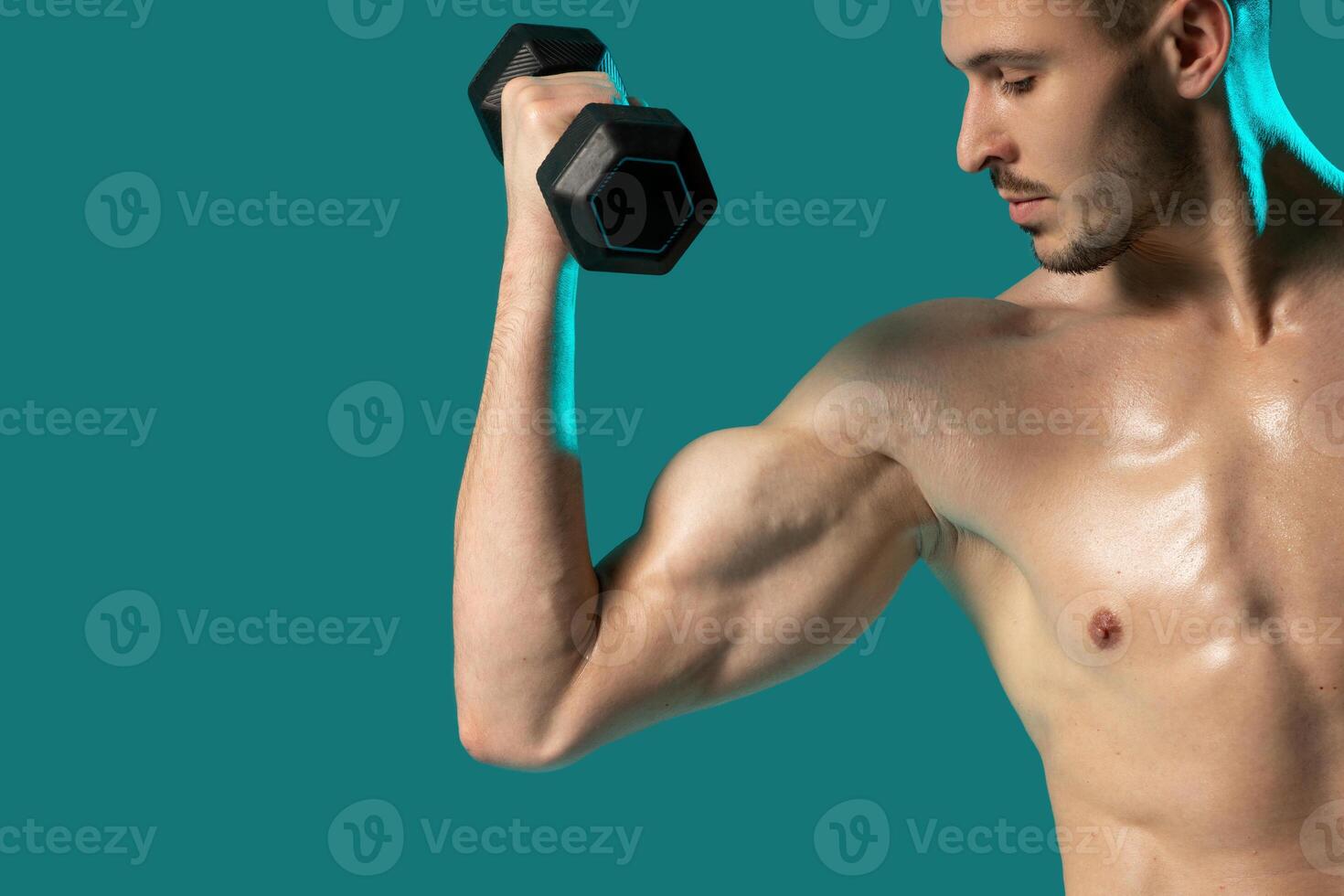 A man is standing in a gym setting, holding a pair of dumbbells and posing for the camera. The background is a solid blue color, emphasizing the man and his workout equipment. photo