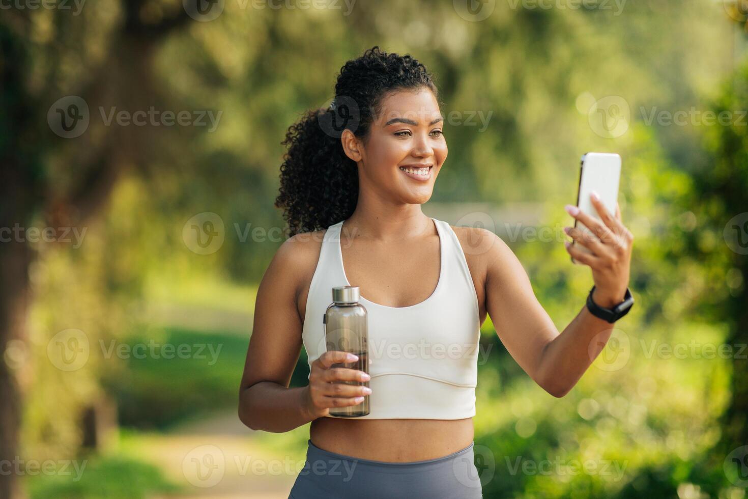 A cheerful young woman is enjoying her post-workout routine in a lush park. She holds a water bottle in one hand while capturing a selfie with her smartphone. photo