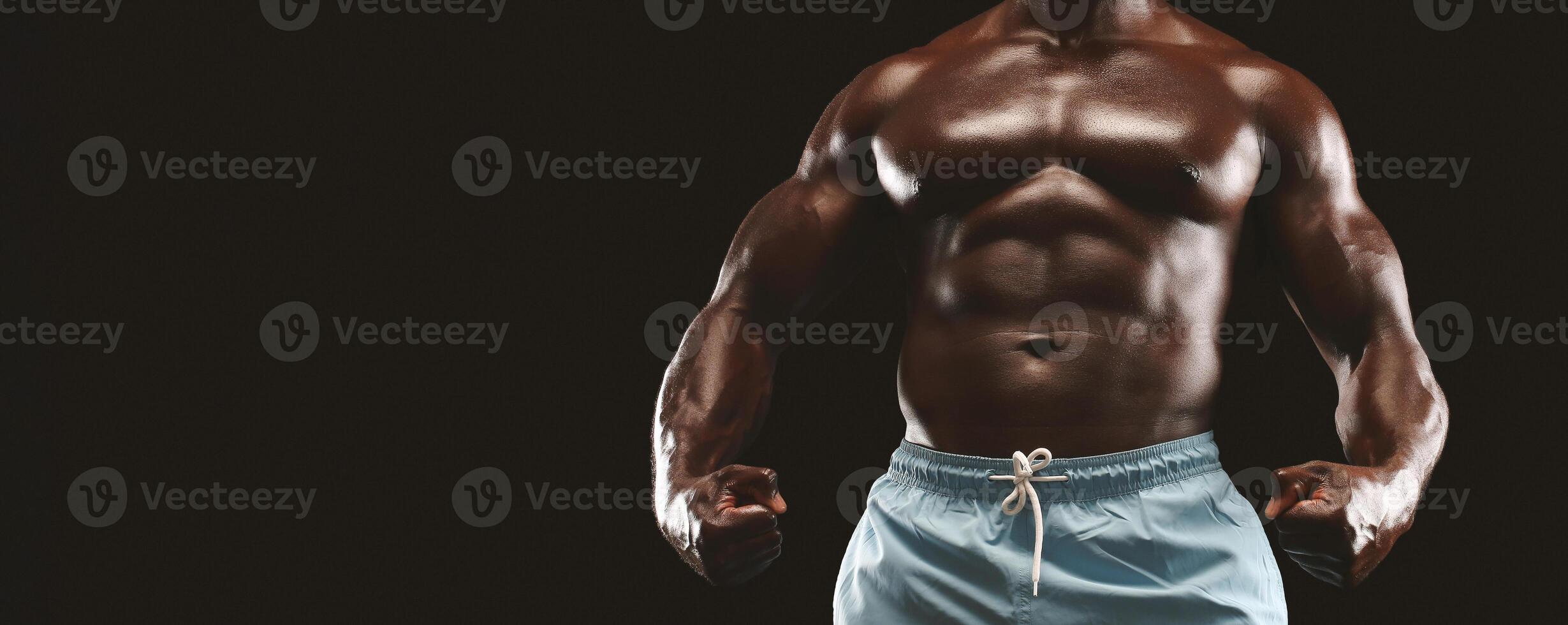 A muscular man with a toned physique poses with clenched fists in front of a black background. He is wearing blue shorts and appears to be in excellent shape. photo