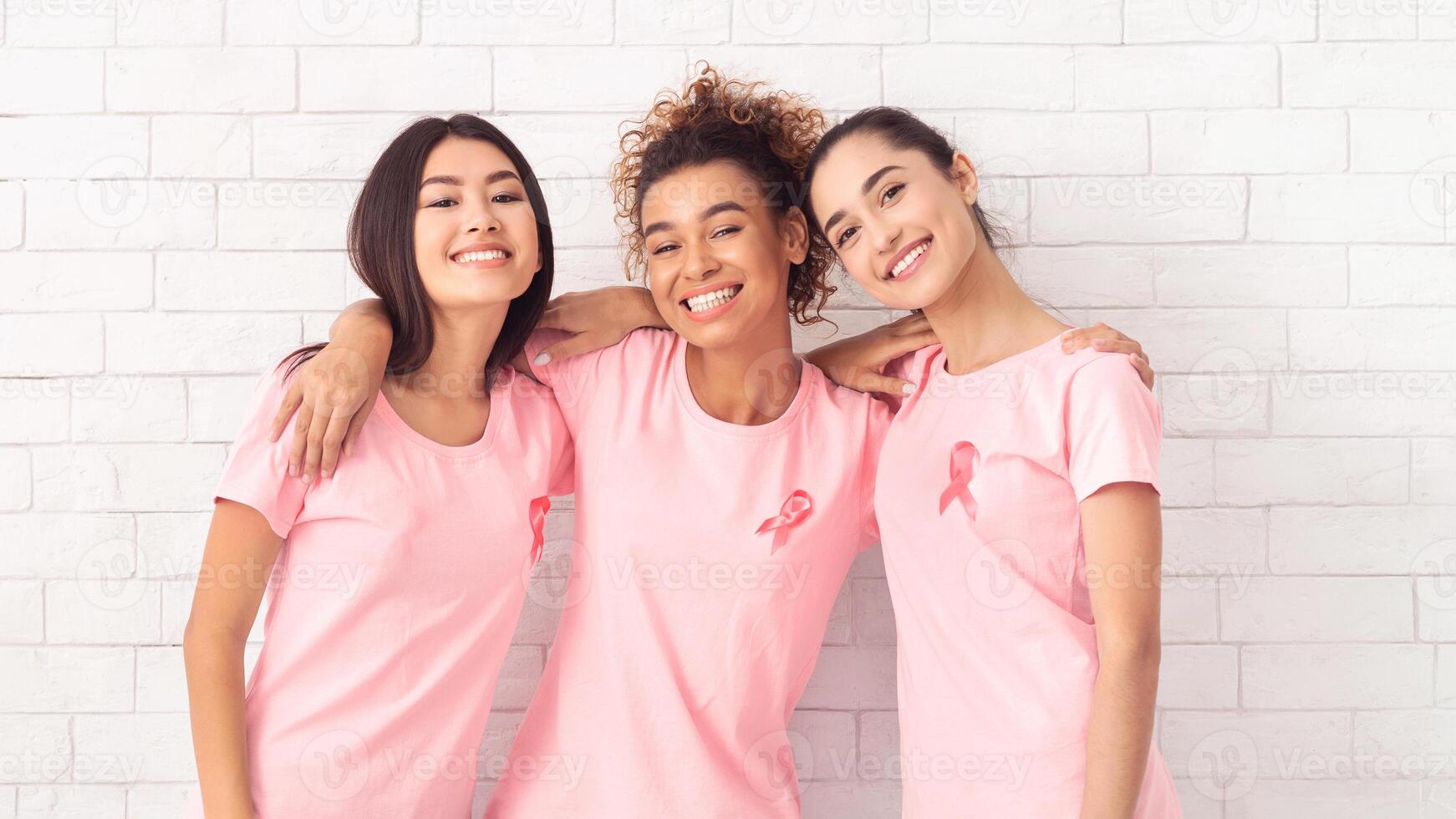 Oncology Support Group. Cheerful Multiracial Women In Pink Breast Cancer T-Shirts Embracing Standing Near Brick Wall. photo