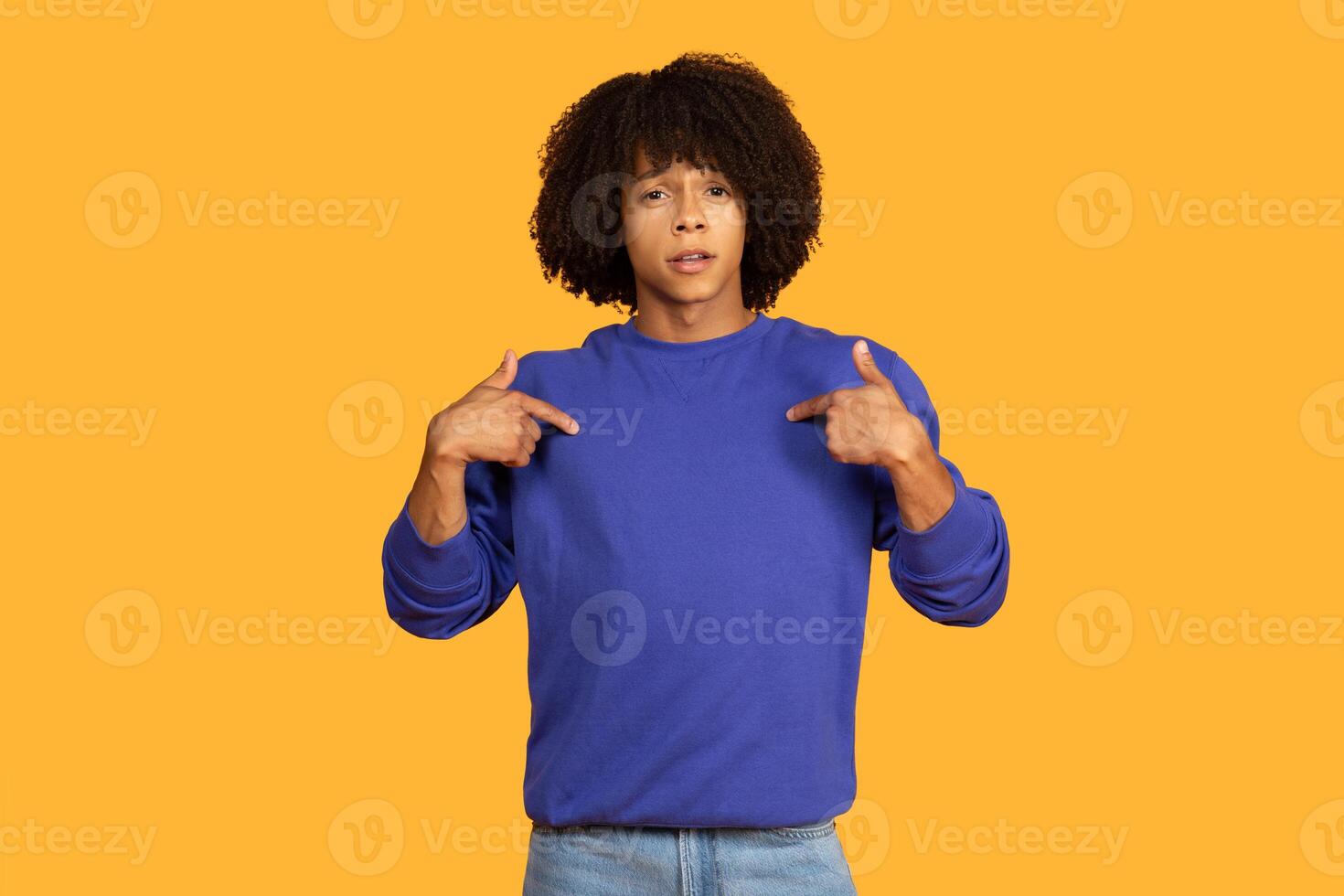A young man wearing a blue pullover is seen pointing towards himself while standing against a vibrant yellow background. His facial expression suggests he is emphasizing or making a point. photo
