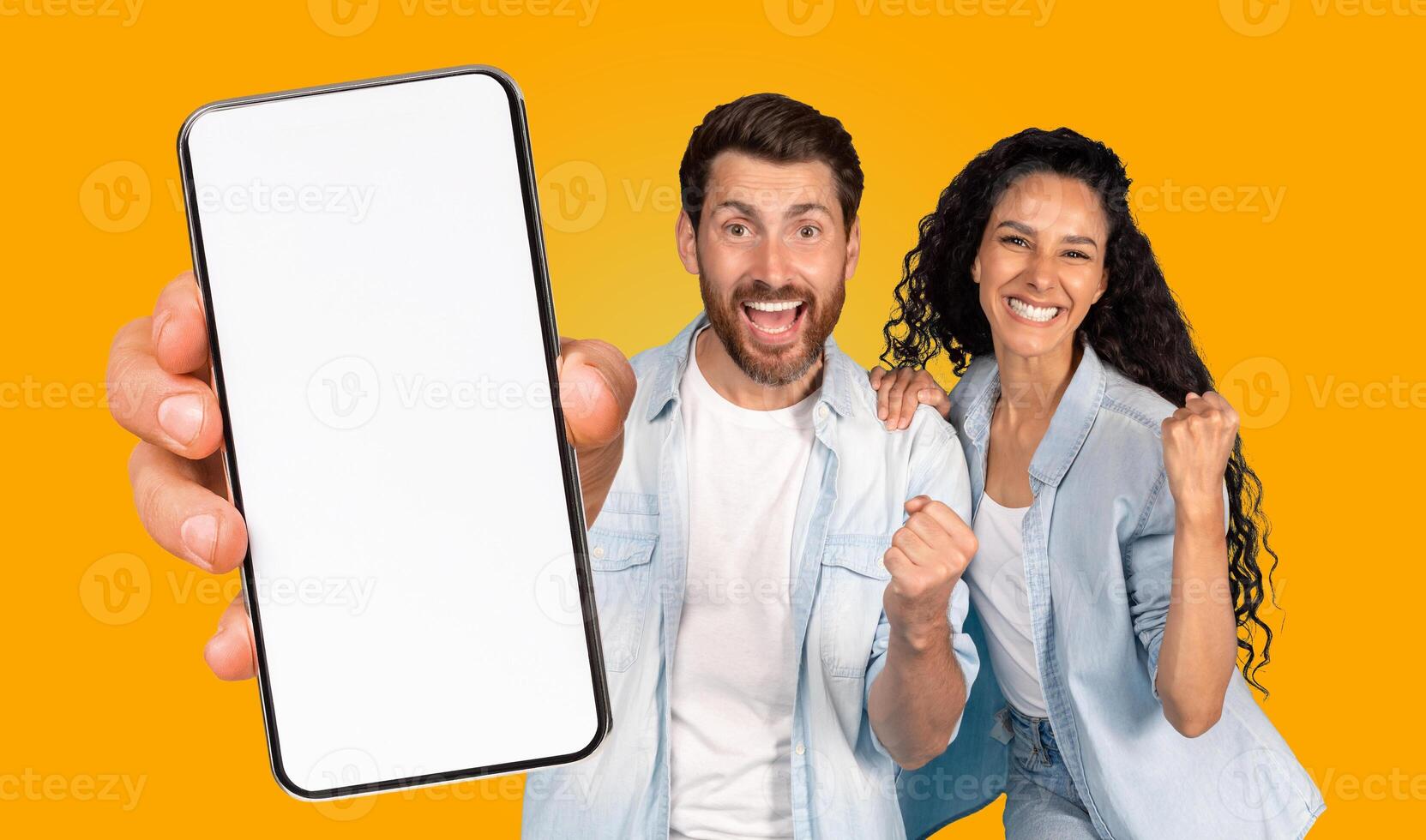 A man and woman stand shoulder-to-shoulder against a vibrant yellow backdrop. The man is holding a smartphone in front of him. The woman raising her right fist in the air photo
