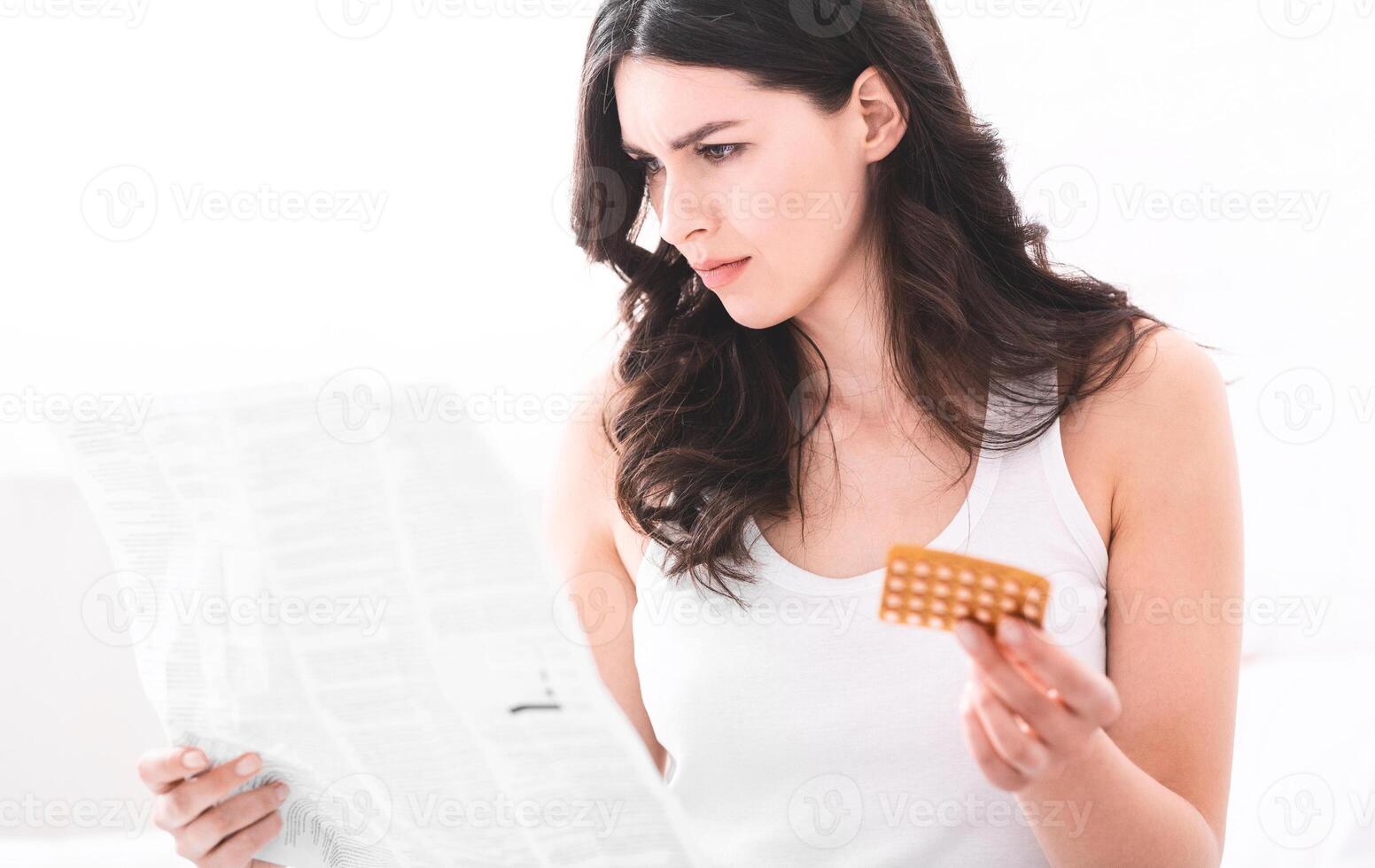 A young woman with long dark hair sits in bed, holding a sheet of paper in her left hand and a pack of birth control pills in her right hand. She is looking down at the instructions on the paper photo