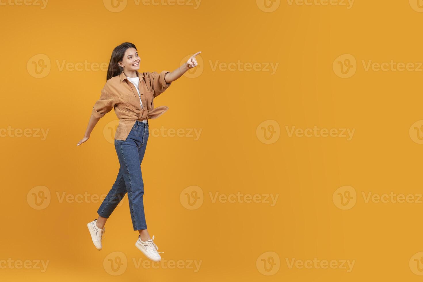 This image shows a woman in a brown shirt and blue jeans jumping in the air while pointing her right hand to the left side of the frame, against a yellow background, copy space photo