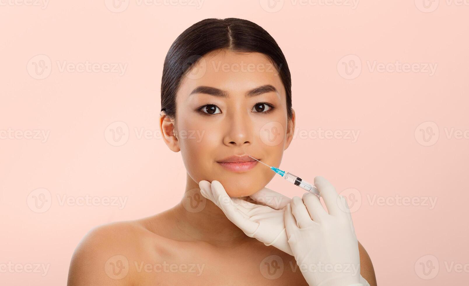 A young Asian woman receives a lip injection from a medical professional. The woman has dark hair pulled back, light skin, and is looking directly at the camera photo