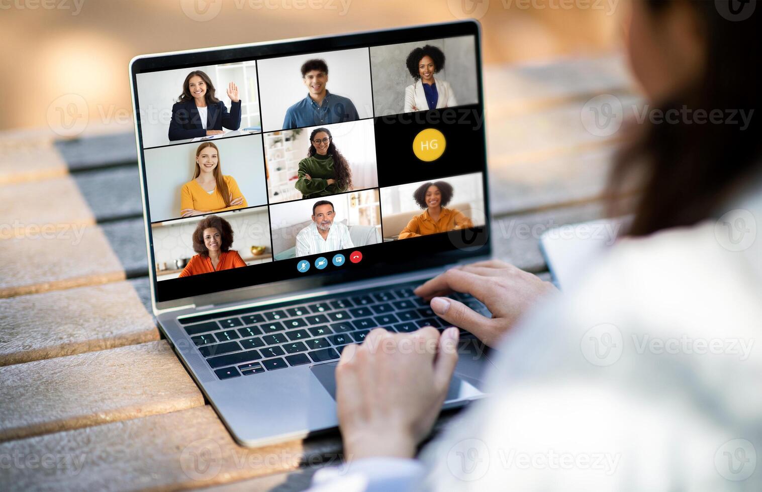 Unrecognizable woman sitting at a desk, typing on a laptop while conferencing with a diverse group of people displayed on the screen. The individuals on screen are engaged in a virtual meeting photo