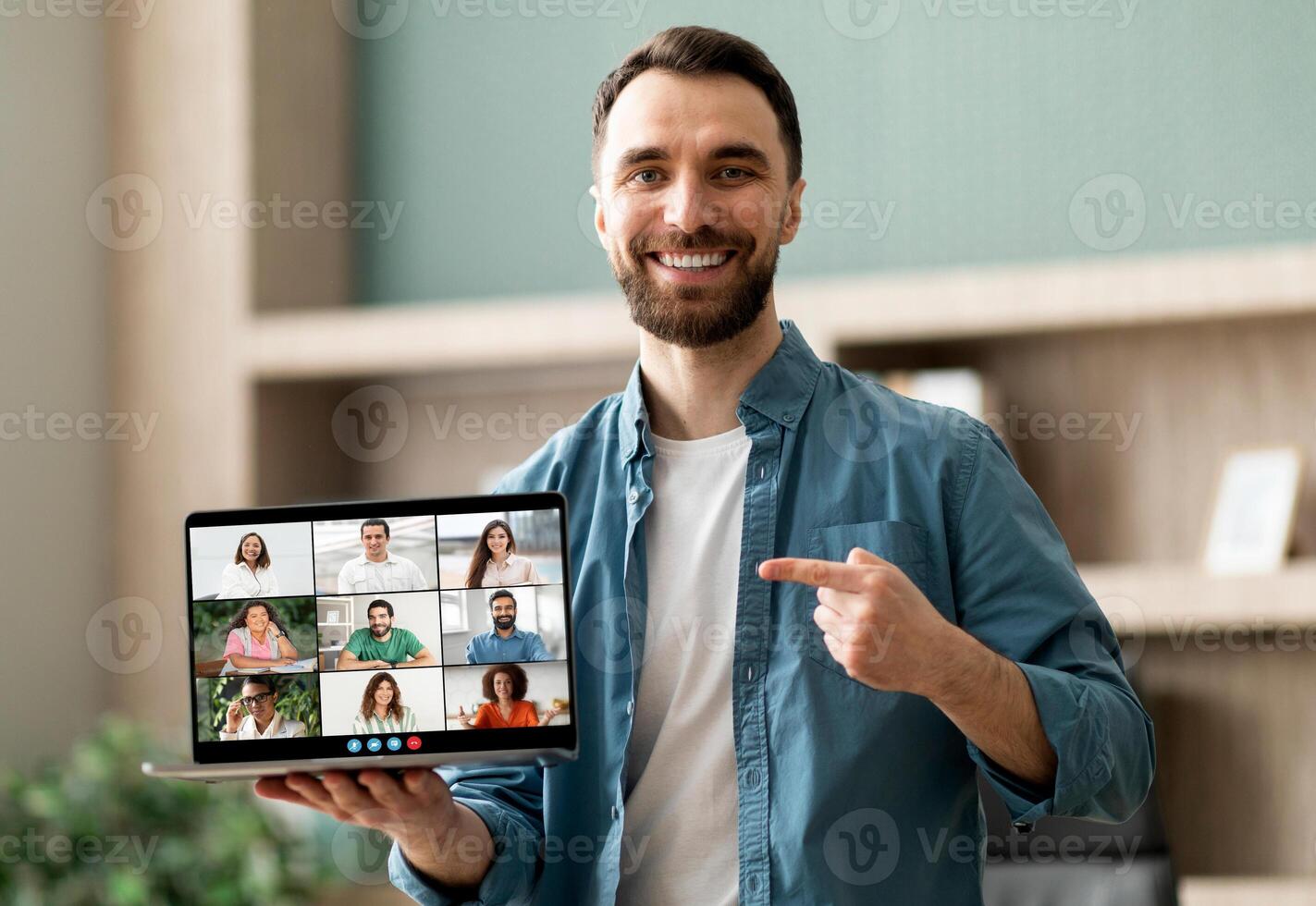 A man is holding a laptop displaying a group of people. He is engaging with the content on the screen while the individuals on the tablet appear focused and engaged as well. photo
