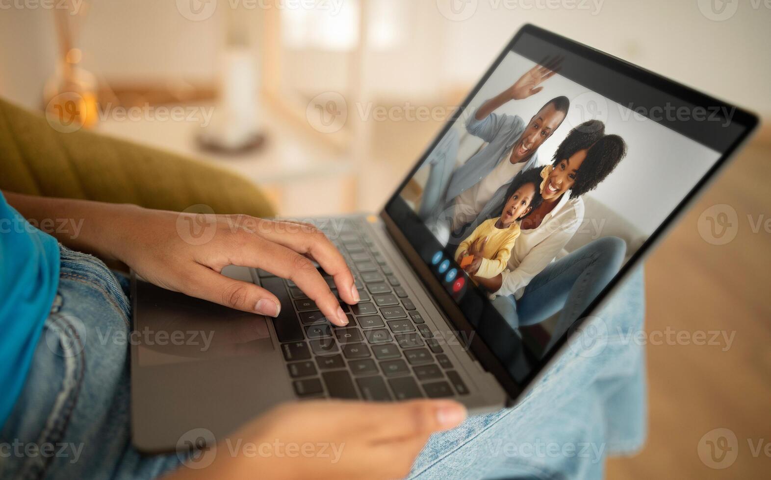 Black lady sits on a couch, using a laptop computer to chat with a family of three. The family on the screen consists of a man, a woman, and a young child, all smiling and waving. photo