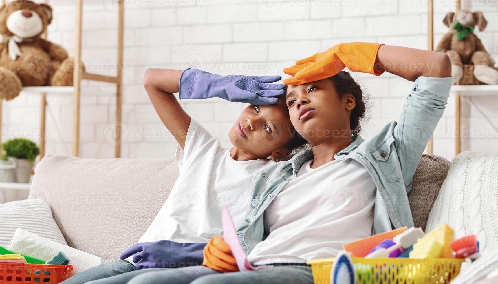 Tired black sisters with cleaning gloves on sitting on sofa, having break while cleaning, empty space photo