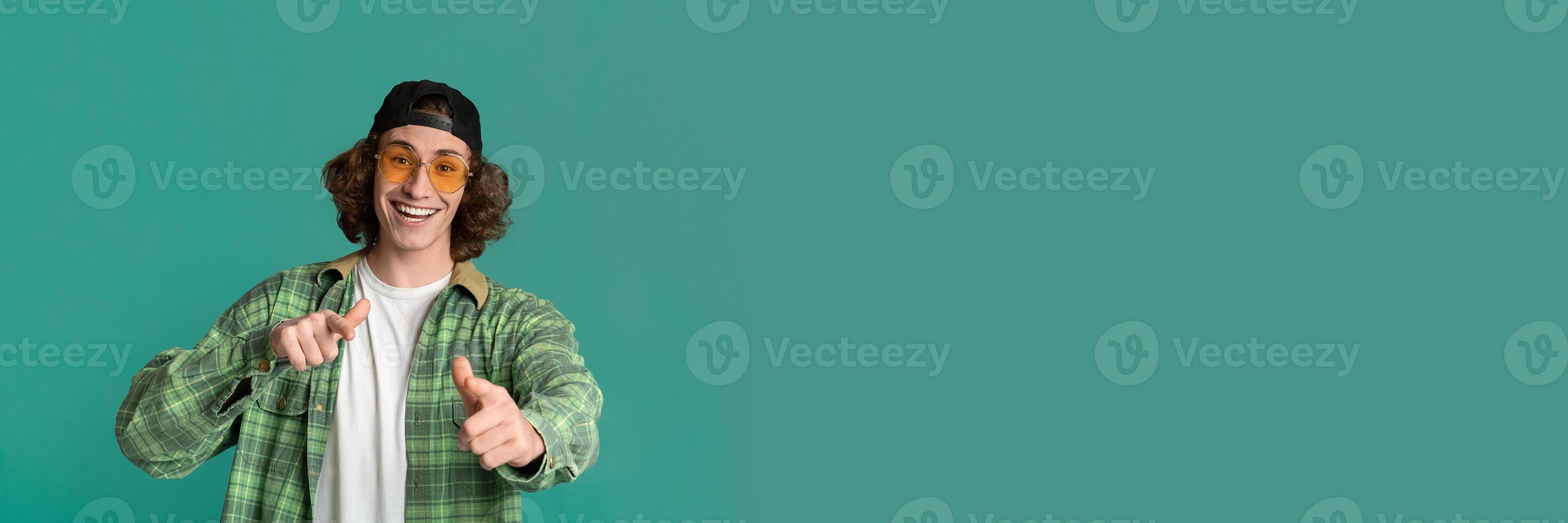 Portrait of cool young guy in sunglasses pointing at you on turquoise studio background, panorama with copy space photo