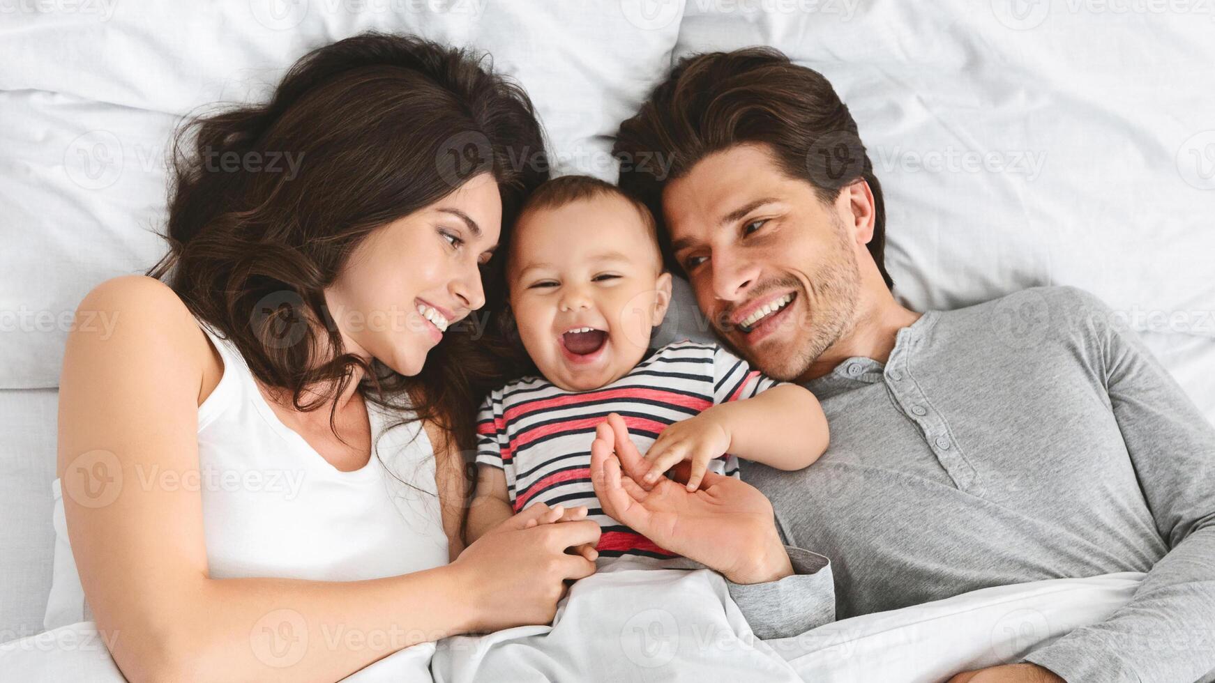 This image shows a happy family of three in bed together during the day. The parents are smiling and looking at their baby, who is laughing and reaching out to touch his fathers face. photo