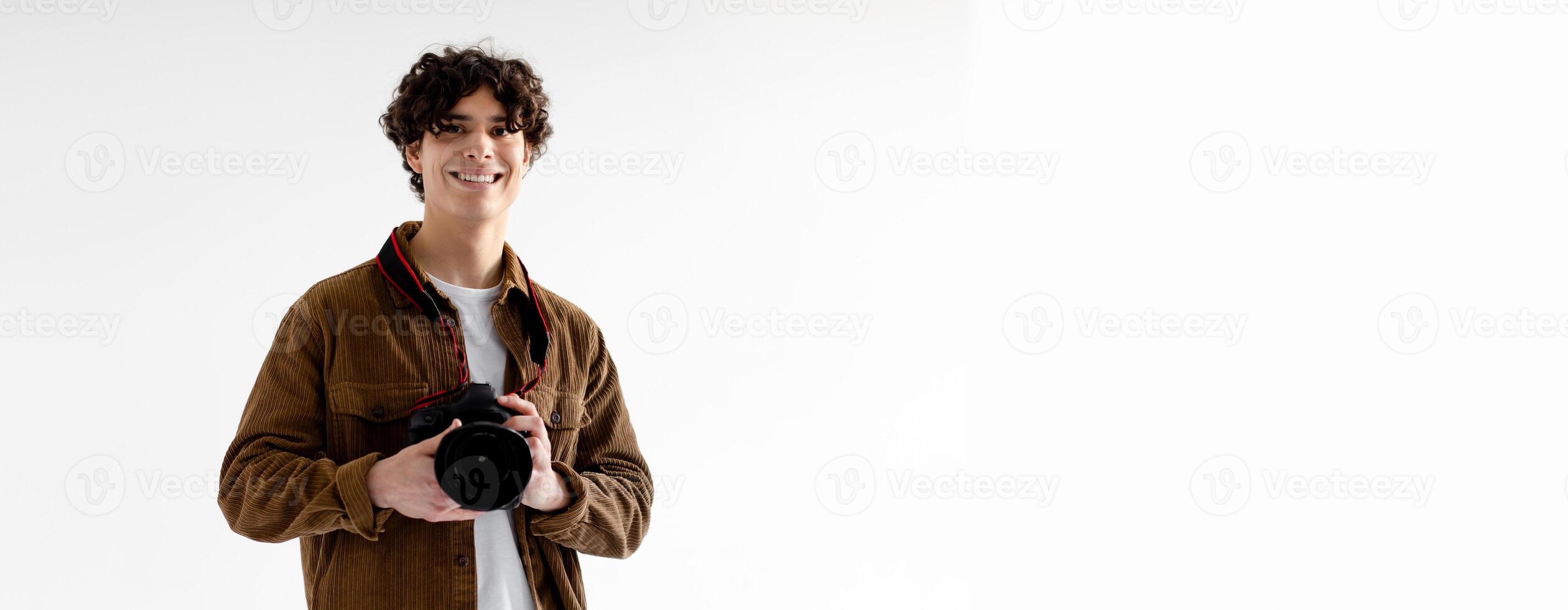A young guy with curly hair stands confidently in a minimalist studio, smiling broadly while holding a camera. The bright, neutral background emphasizes their enthusiasm and passion for photography photo