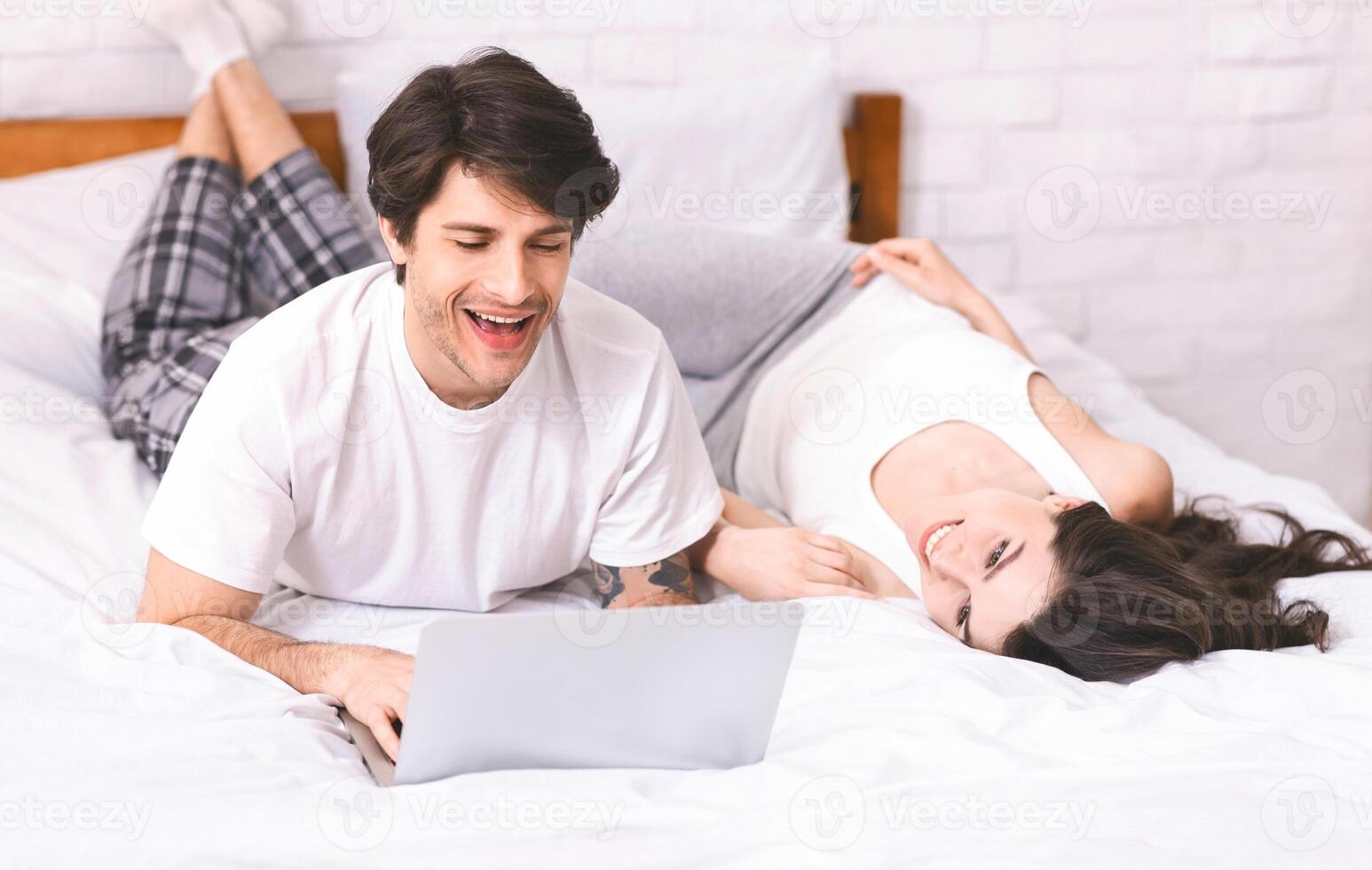 A man and a woman are relaxing in bed together. The man is using a laptop and smiling while looking at the screen. The woman is lying on her back with her head turned to look at him, smiling. photo