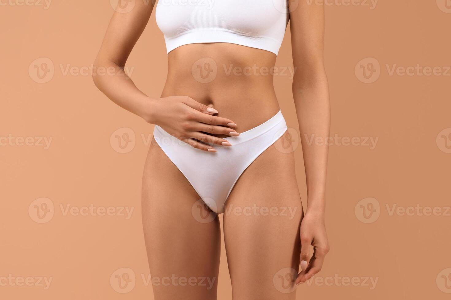 A woman is wearing white underwear and has her hands resting on her stomach. The background is a muted orange color, cropped photo