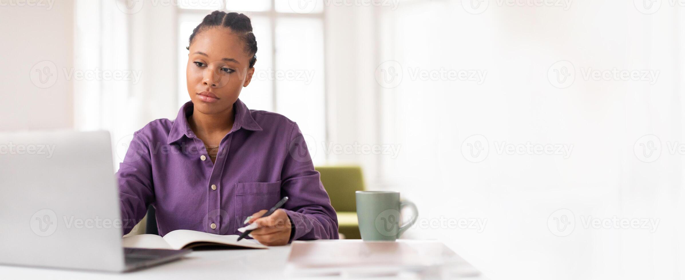 A young black woman focused on her studies at a bright and cozy workspace. She takes notes while using her laptop and enjoys a warm beverage, creating a productive atmosphere in the afternoon light. photo