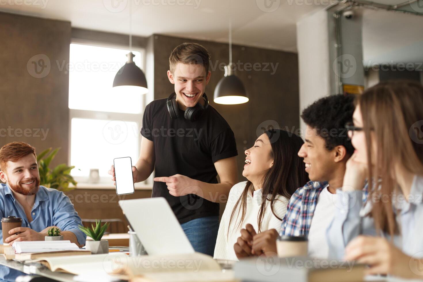 A group of young adults is gathered around a table in a well-lit co-working space, engaging in a lively discussion. One man stands showing his smartphone to the group photo