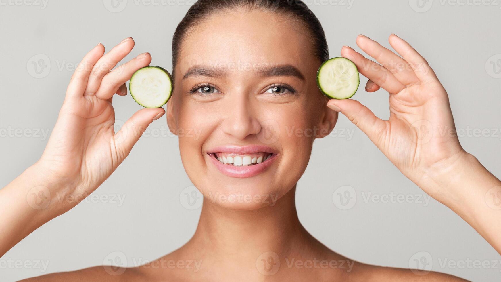 A young woman with a bright smile holds cucumber slices near her eyes against a plain background. She showcases a fresh and healthy skincare routine. photo