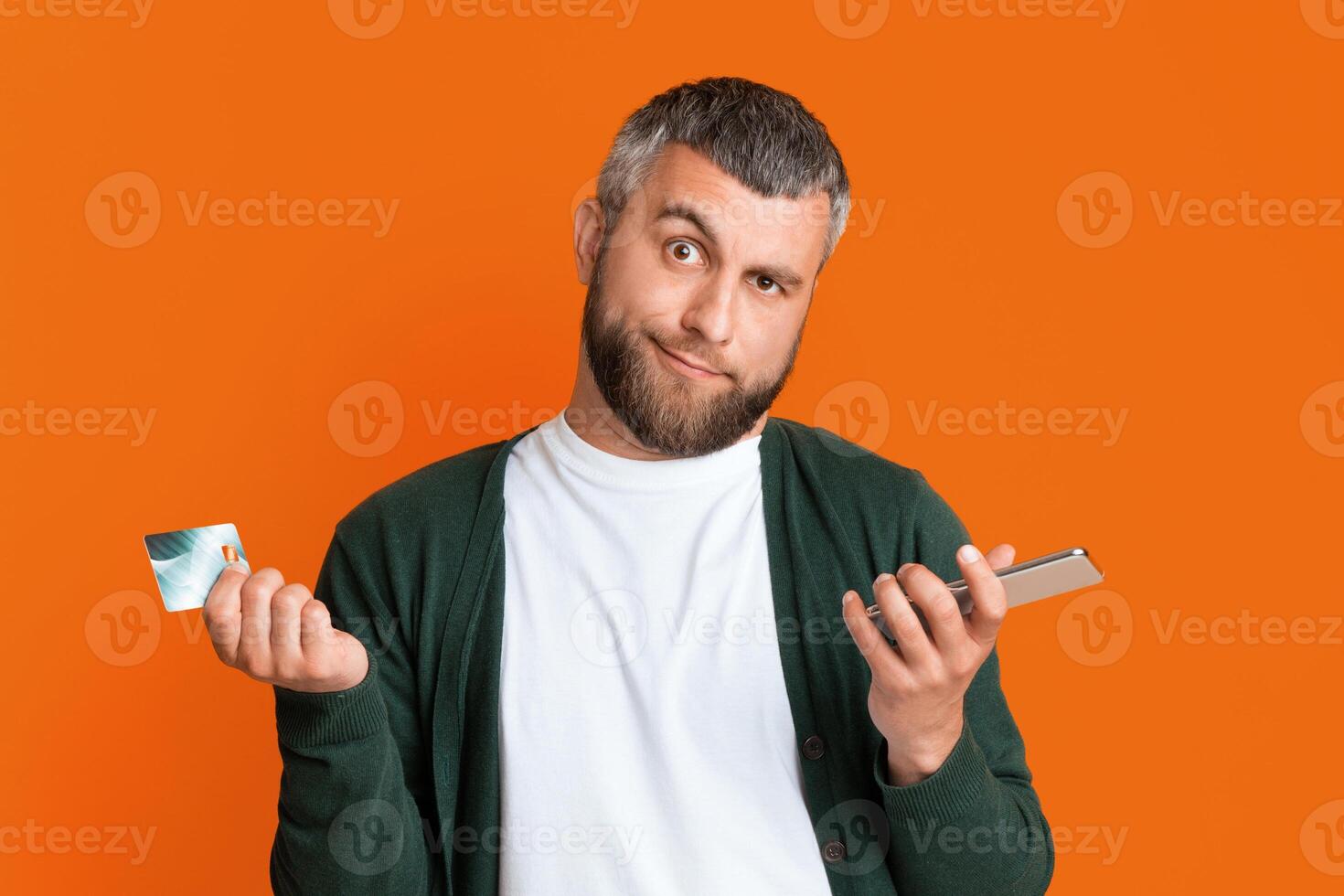 Confused bearded man holding smartphone and credit card in his hands and grimacing, isolated on orange studio background photo
