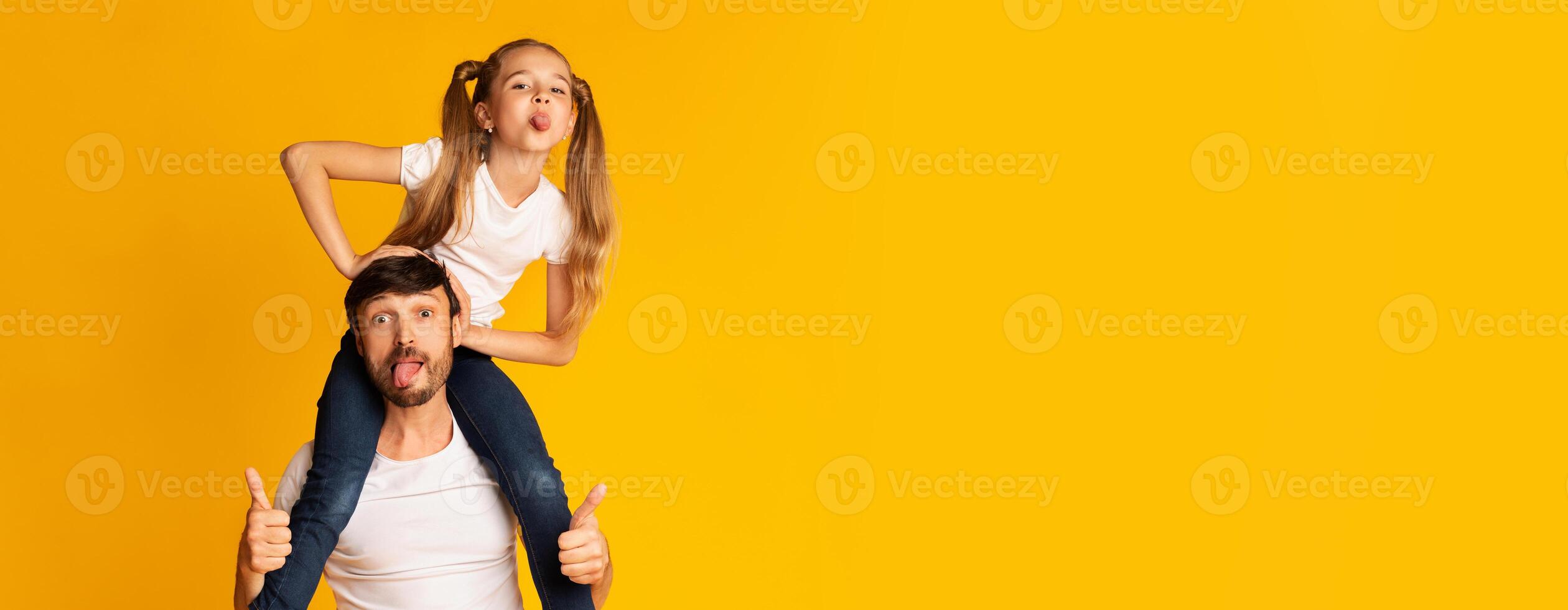 A father gives his daughter a fun ride on his shoulders while both make playful faces, capturing a joyful moment of their bond and laughter. Vibrant yellow backdrop enhances the cheerful atmosphere. photo