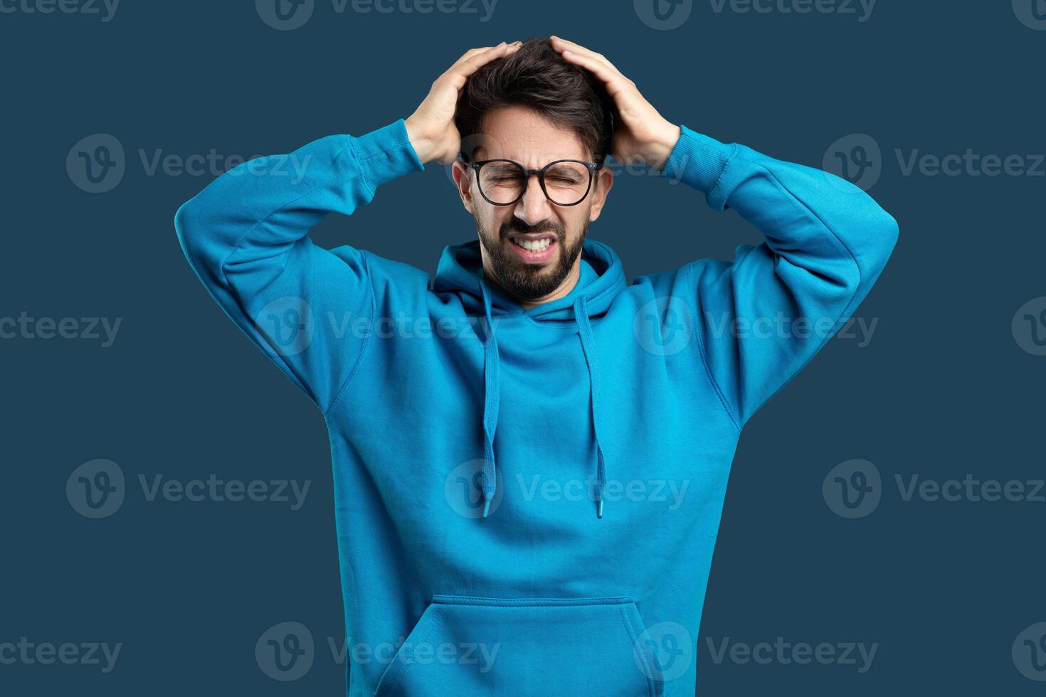 A young man with a beard and glasses, wearing a blue hoodie, is standing against a solid blue background. He appears frustrated or stressed photo