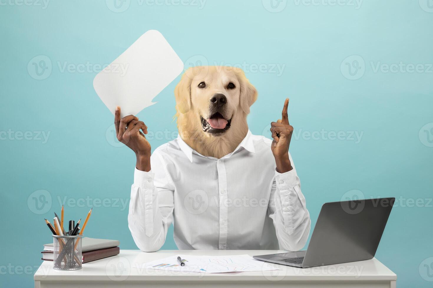 A person with the head of a golden retriever raises one hand while holding a blank speech bubble with their other hand. They are sitting at a desk with a laptop and stationery photo