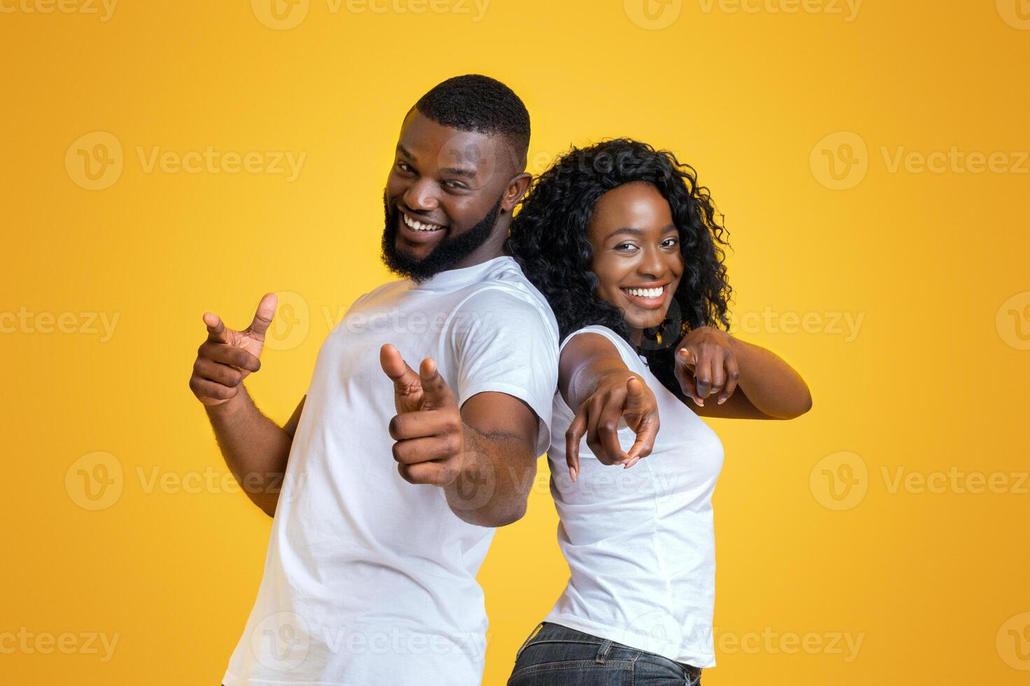 Cheerful African American Man And Woman Pointing Fingers At Camera, staying back to back on yellow studio background, free space photo