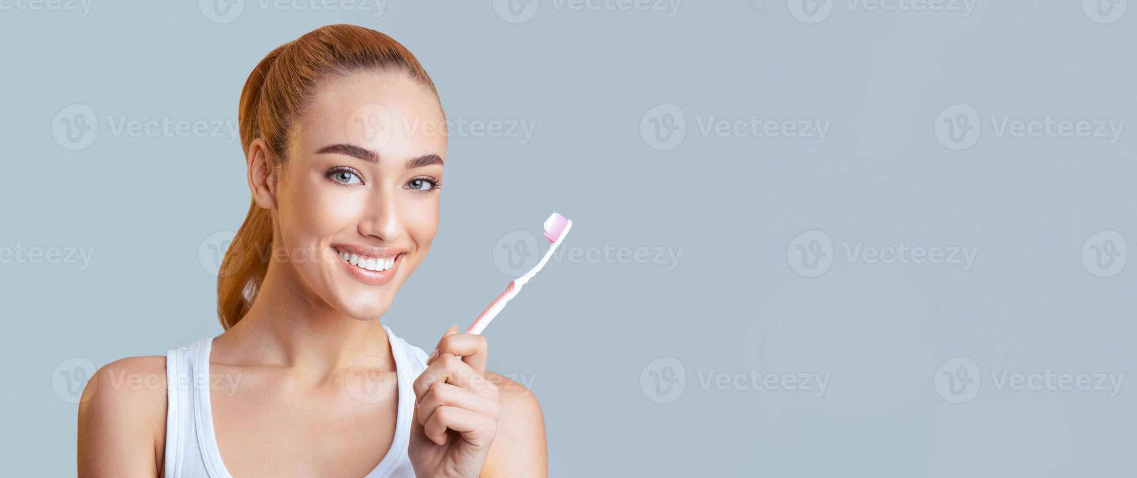 A young woman with red hair and a bright smile is holding a pink toothbrush. She is ready to brush her teeth and keep her oral health in check. She is looking directly at the camera, panorama photo