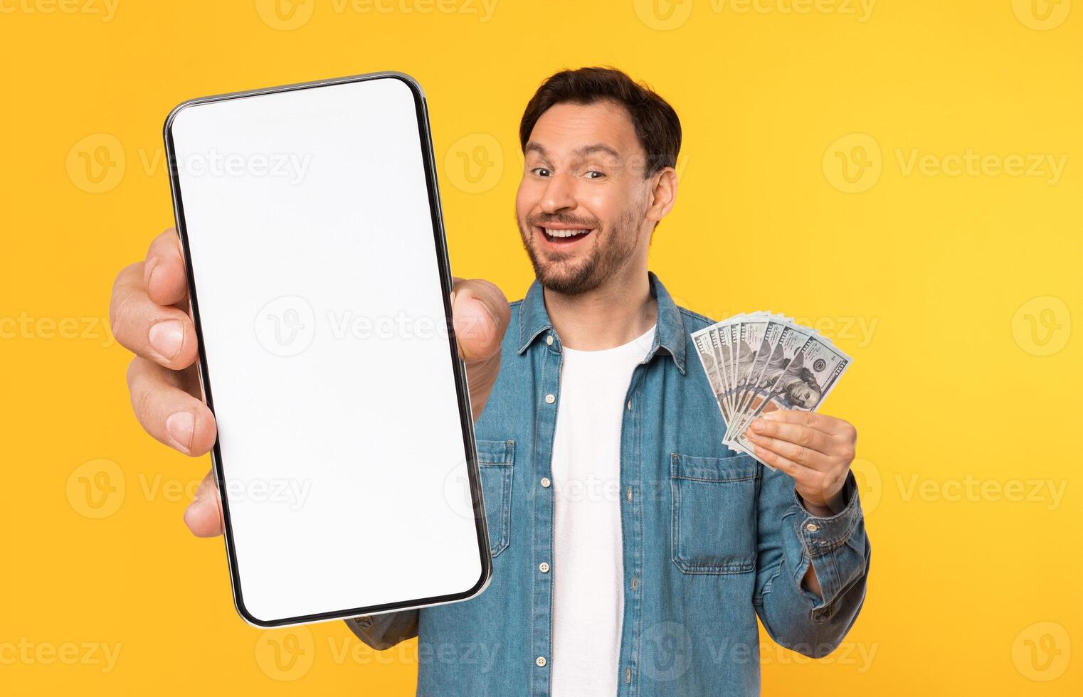A smiling man displays a blank smartphone in one hand while holding a stack of cash in the other. The vibrant yellow background enhances the cheerful atmosphere of the moment. photo