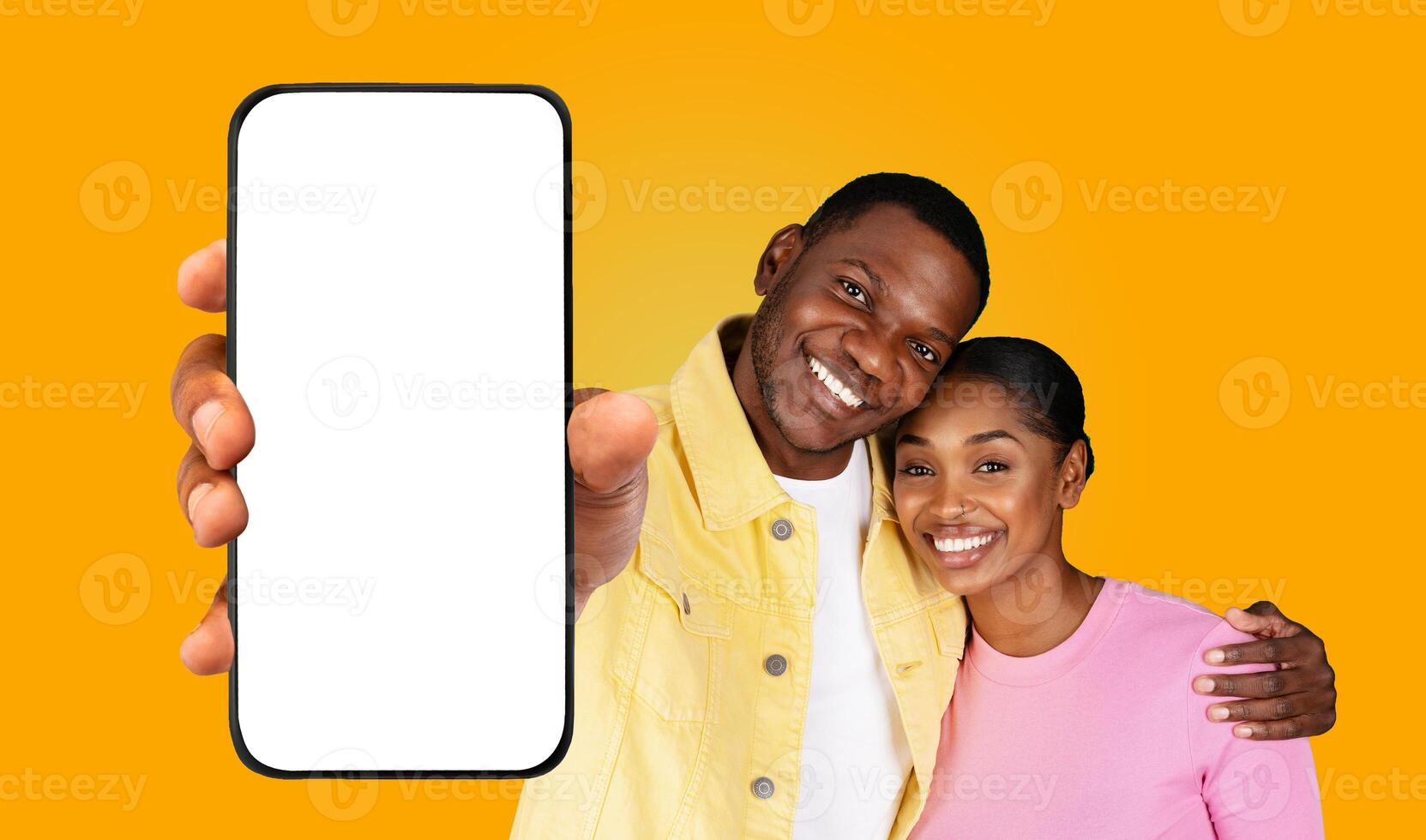 A smiling black couple stands together in front of a yellow background, with the man holding a smartphone in front of him. The woman is leaning on the man's shoulder, and they both look happy photo