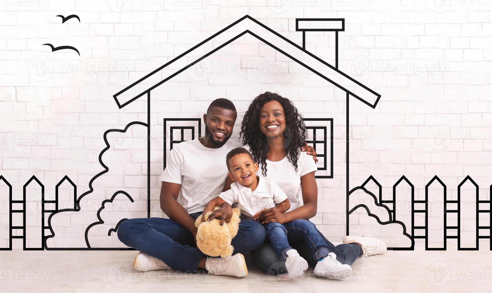 Happy black family of three father mother and little son sitting on floor against illustrated house of their dreams over white wall background photo