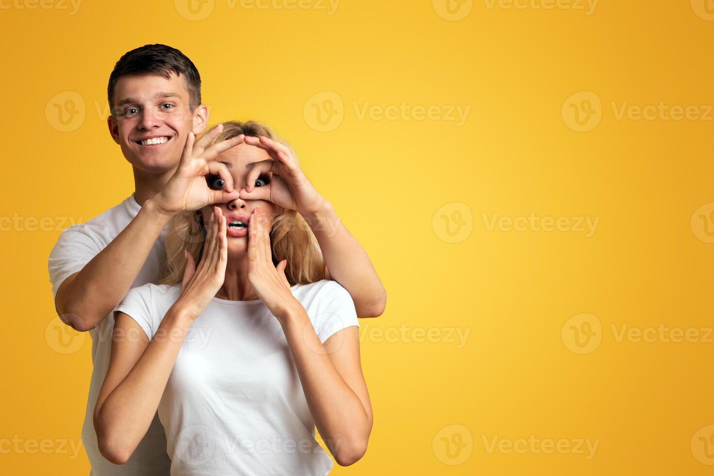 You must see it. Cheerful couple having fun, young guy making funny glasses with fingers to his girlfriend over yellow background with copy space photo