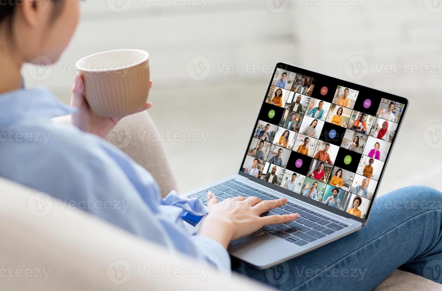 A woman is sitting on a couch, holding a cup of coffee in one hand and using a laptop with a conferencing app open. The screen shows numerous participants in the call. photo
