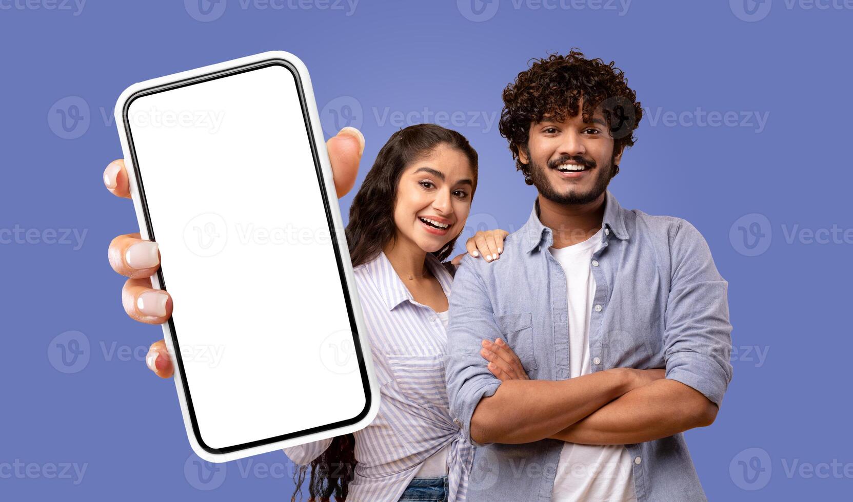 A young Indian couple, stand in front of a solid blue background. The woman is holding a smartphone with a white screen towards the camera, her hand visible in the frame. photo