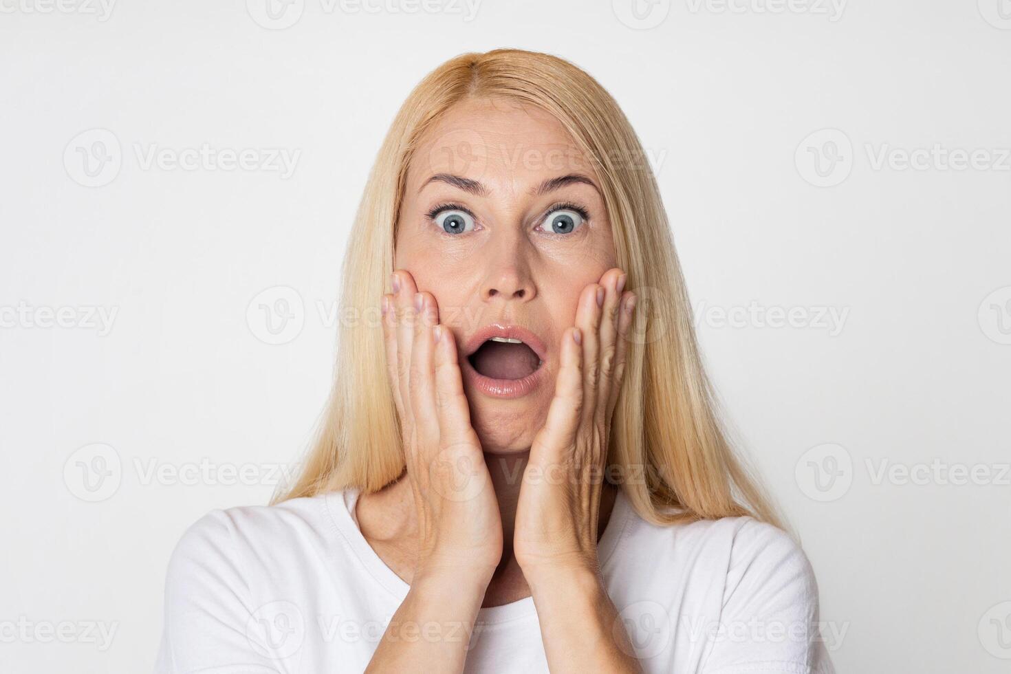 This image shows a woman with blonde hair looking shocked, with her mouth open and her hands covering her cheeks. She is dressed in a white t-shirt and is looking directly at the camera. photo