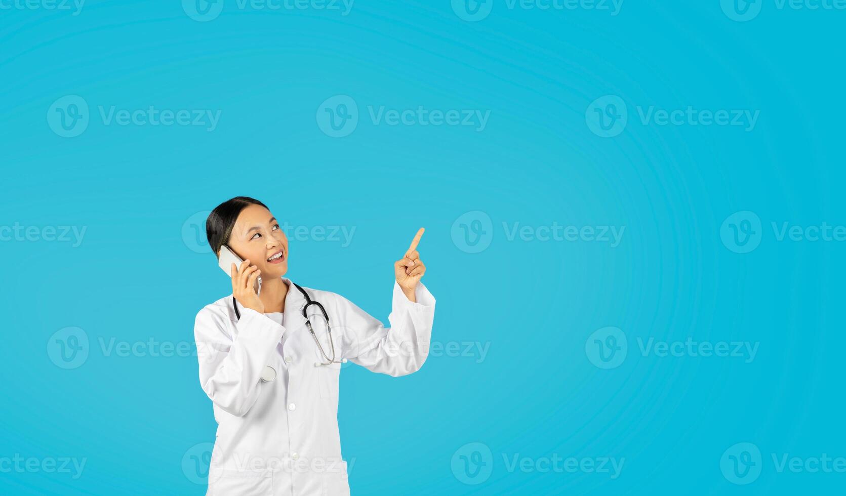 Asian woman doctor in a white coat is on a phone call, smiling and looking up while pointing at copy space. She is wearing a stethoscope around her neck. The background is a solid blue. photo
