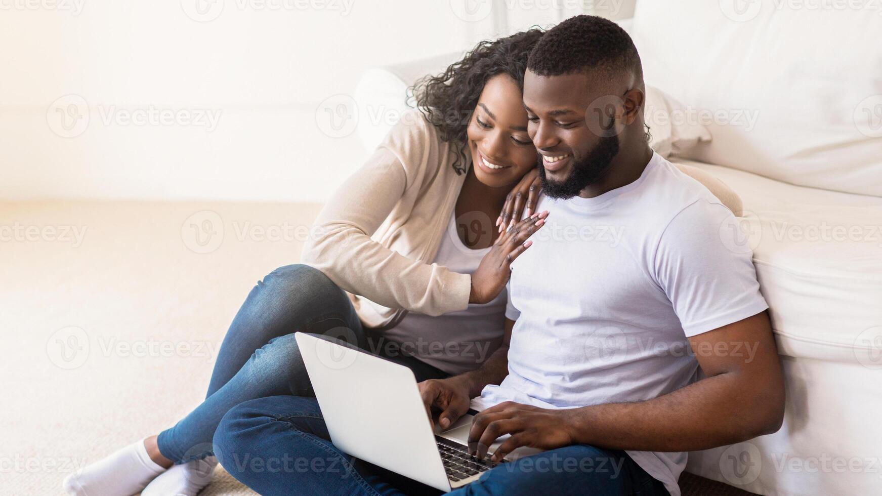 A smiling couple sits on the floor of their living room, using a laptop together. The woman is sitting to the mans left and has her arm around his shoulder. photo