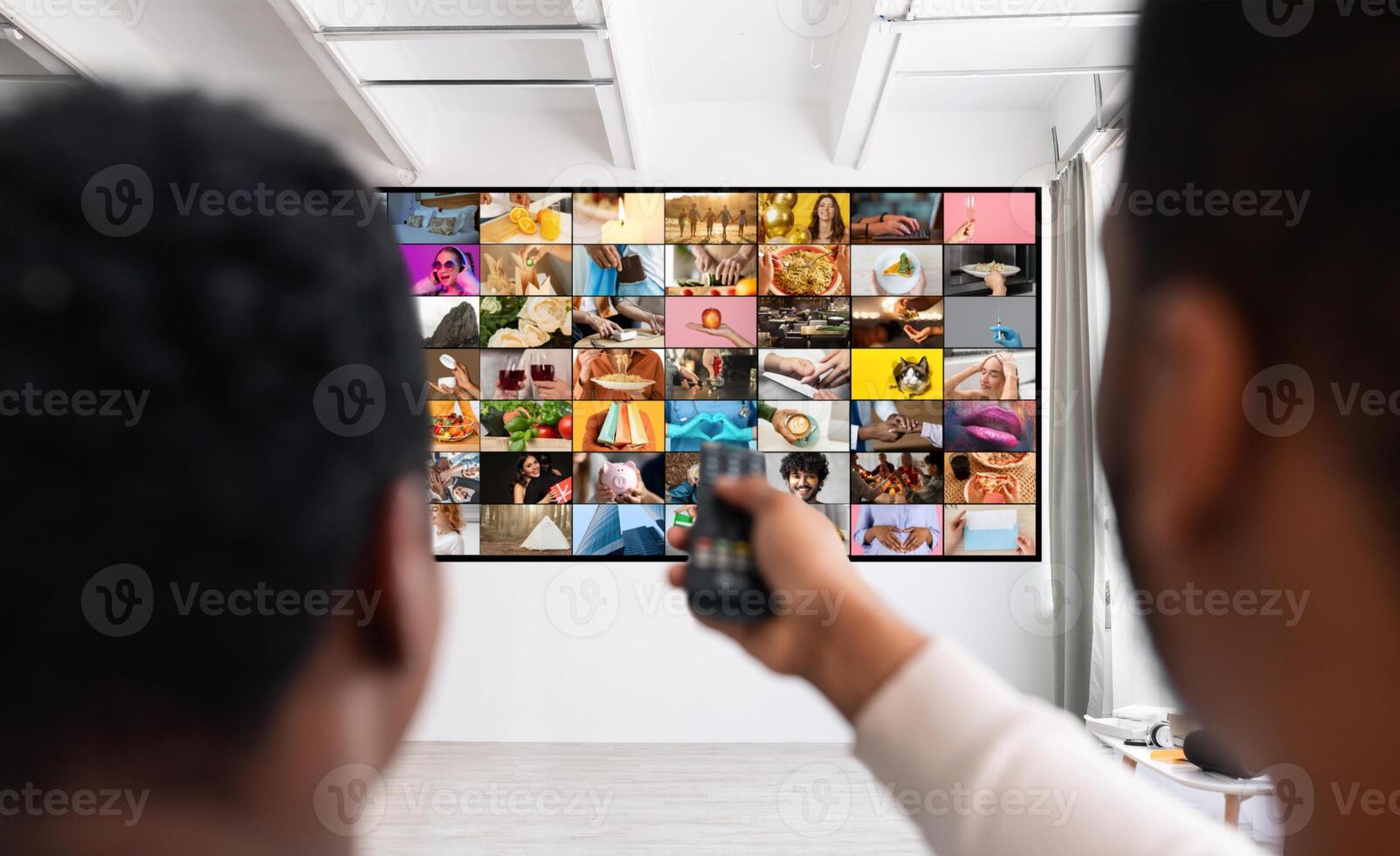 A man is shown from behind, sitting in front of a television displaying a multitude of different channels. He holds a remote control in his hand, presumably changing channels or adjusting the volume photo