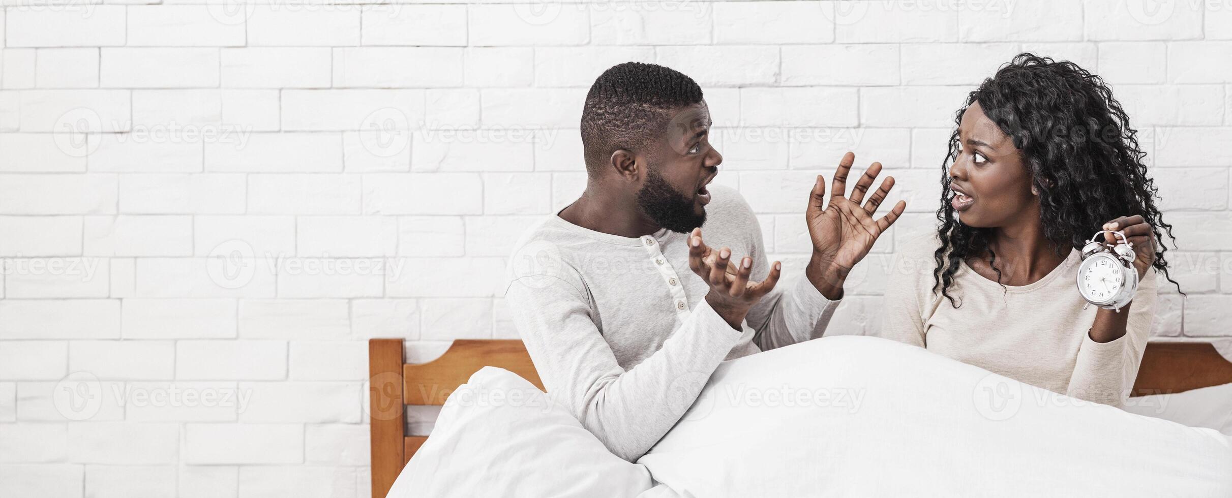 Oh no, we're late. Worried afro couple overslept morning alarm clock, sitting in bed and emotionally reacting, don't know what to do, free space photo