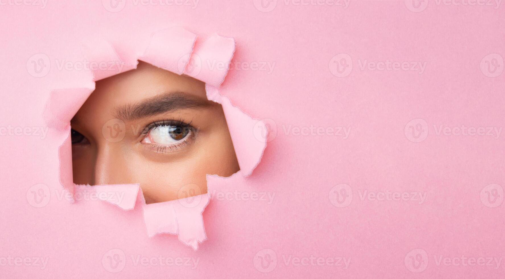 A lady with long brown hair is peeking out of a hole in pink paper. She is looking curious and excited, with her eyes wide open. photo