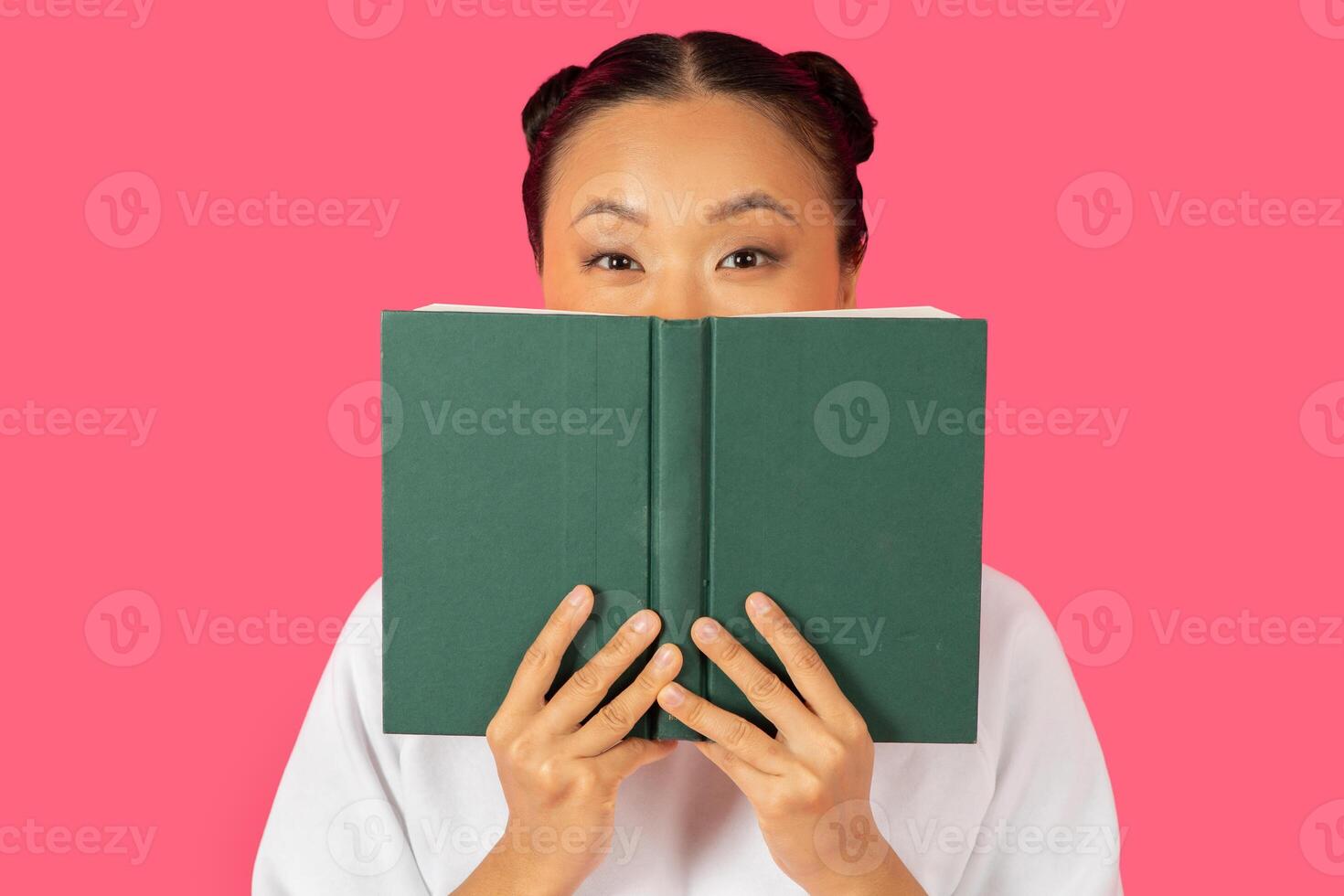 Asian woman joyfully holds a green book in front of her face, with a playful expression, set against a vibrant pink background. photo