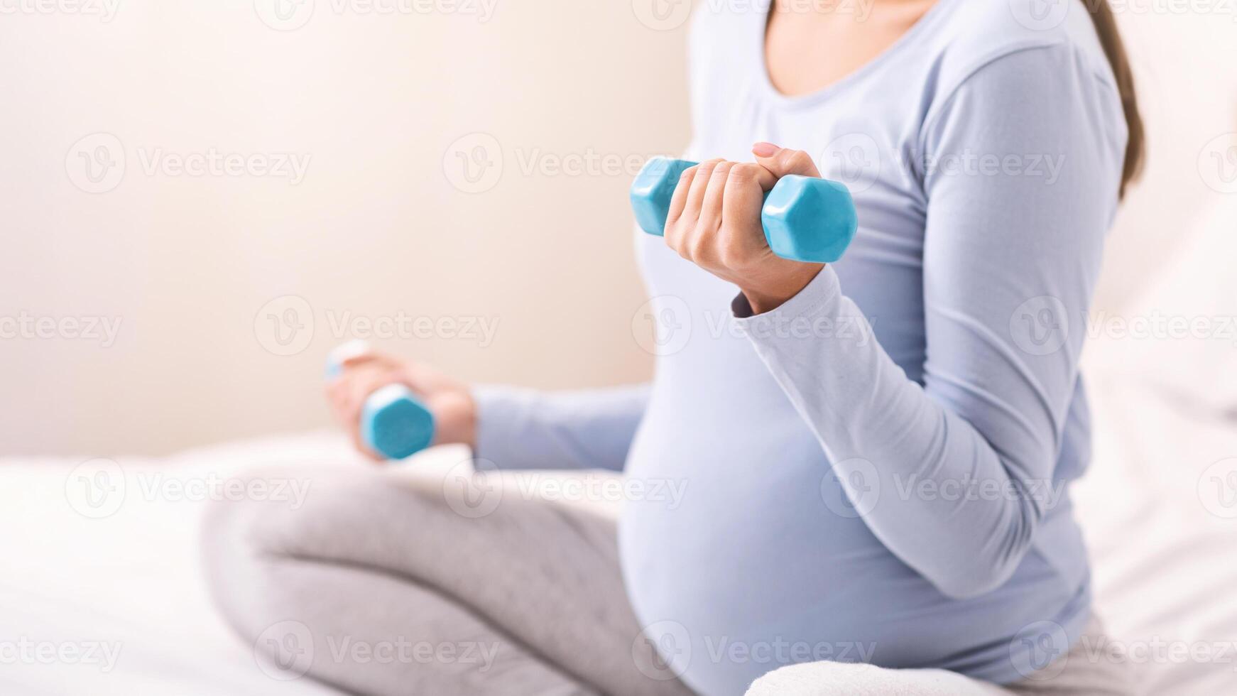 Pregnancy And Sport. Unrecognizable Expectant Young Woman Exercising With Dumbbells Sitting On Bed Indoor. Cropped, Selective Focus photo