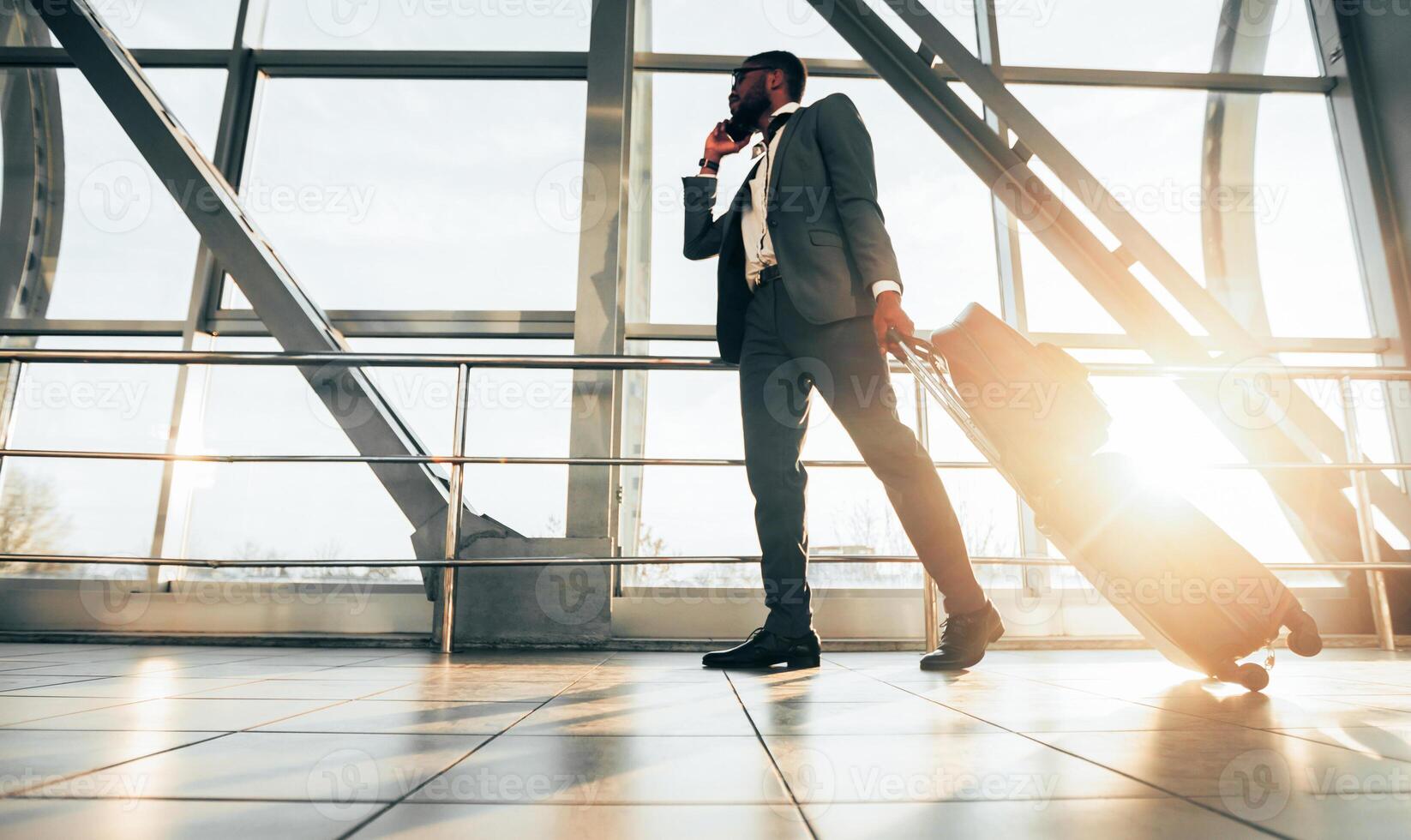 African American man in a suit with luggage making a call outside an airport. The setting is busy and professional, showcasing the multitasking required in business travel. photo