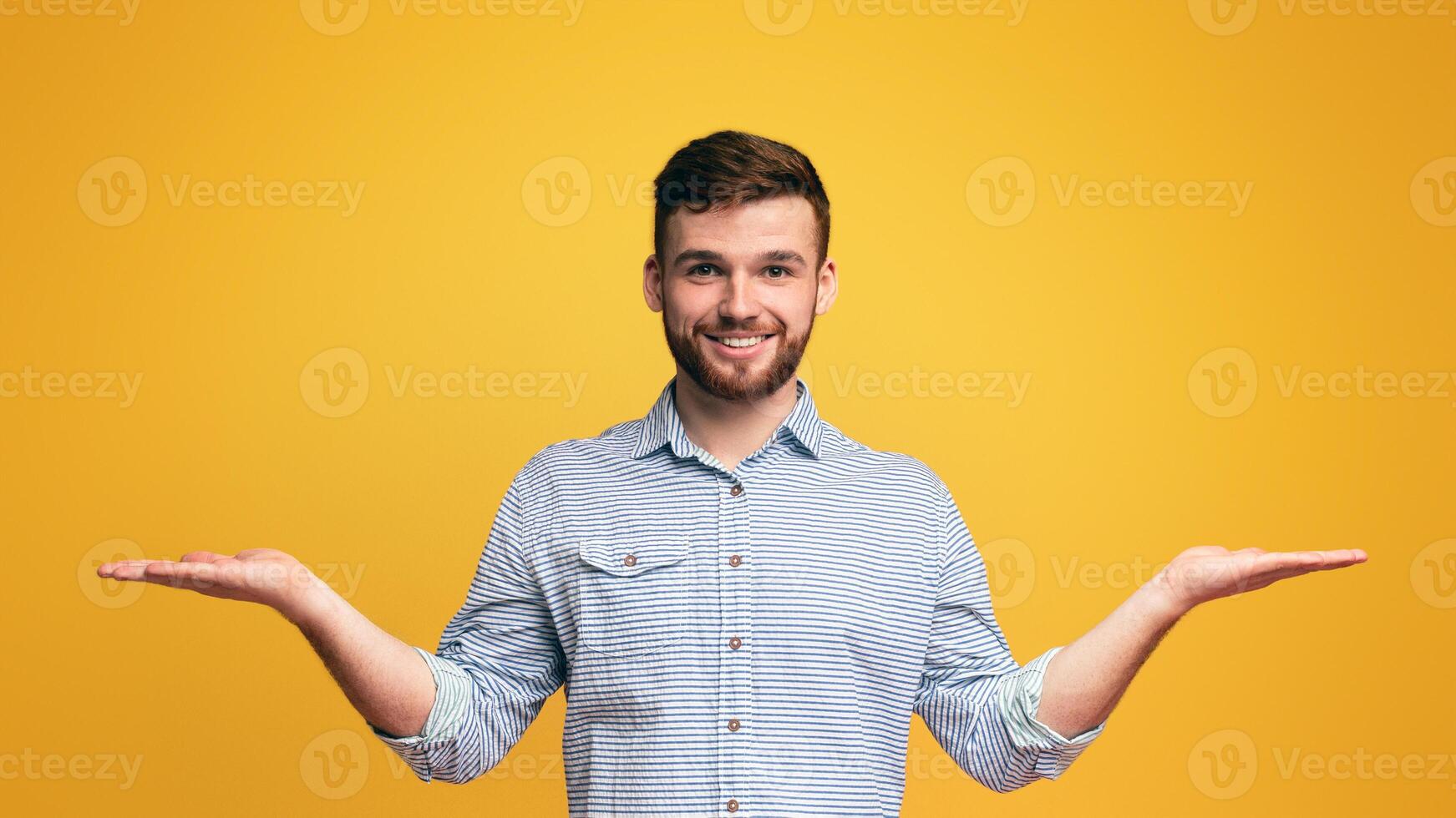 Smiling millennial man holding empty palms up as scales, making choice, copy space, yellow studio background photo