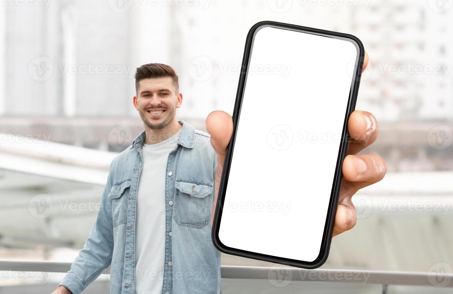 A man is holding a smart phone with white blank screen in his hand. The background is neutral, focusing on the man and the technology in his hand. photo