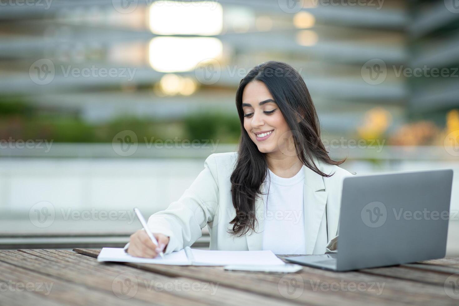 A woman seated at a wooden table enjoys writing notes while using a laptop in an urban setting. photo