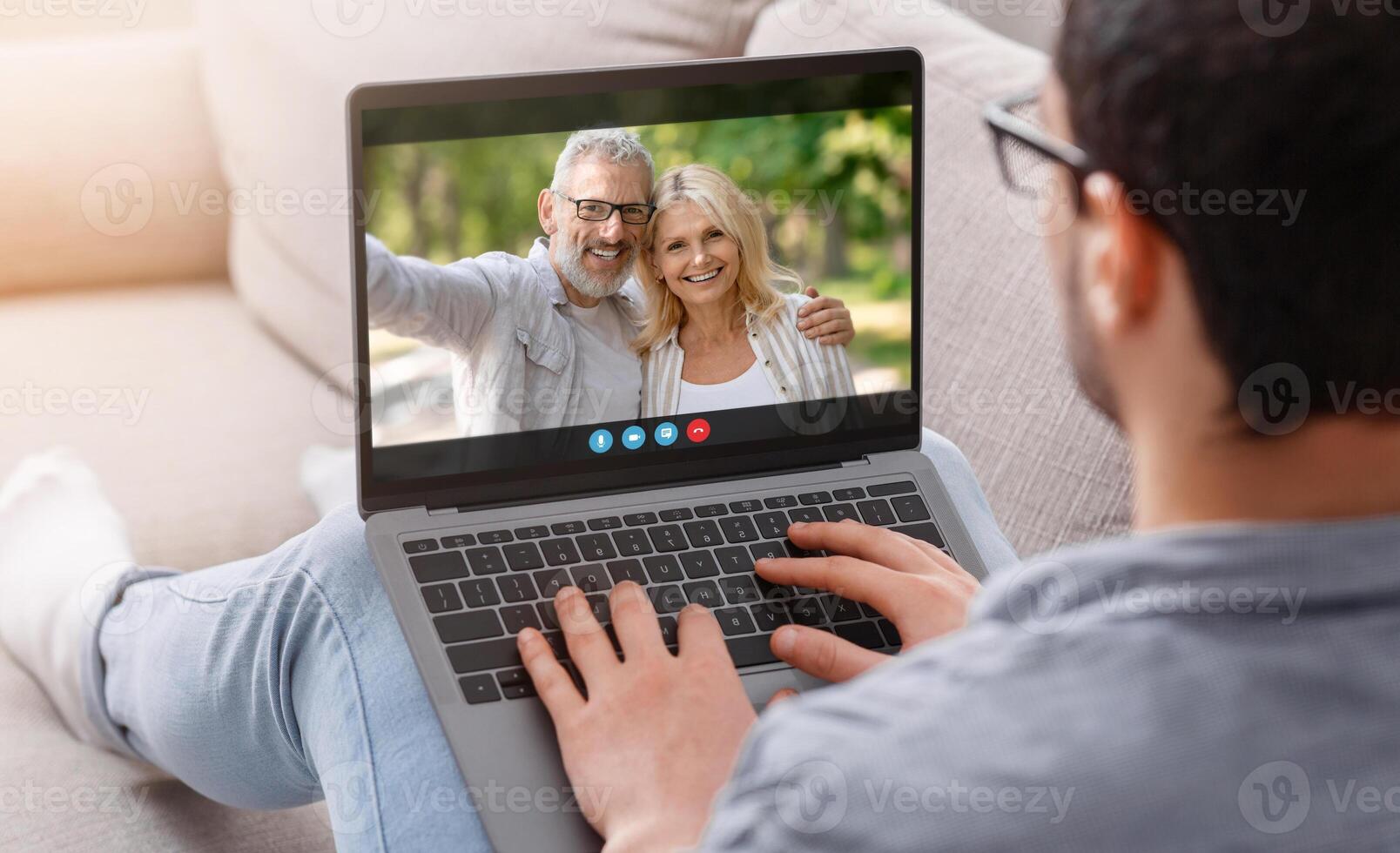 A man is sitting on a couch and chatting with an older couple on his laptop. He is smiling and appears to be engaged in conversation. photo