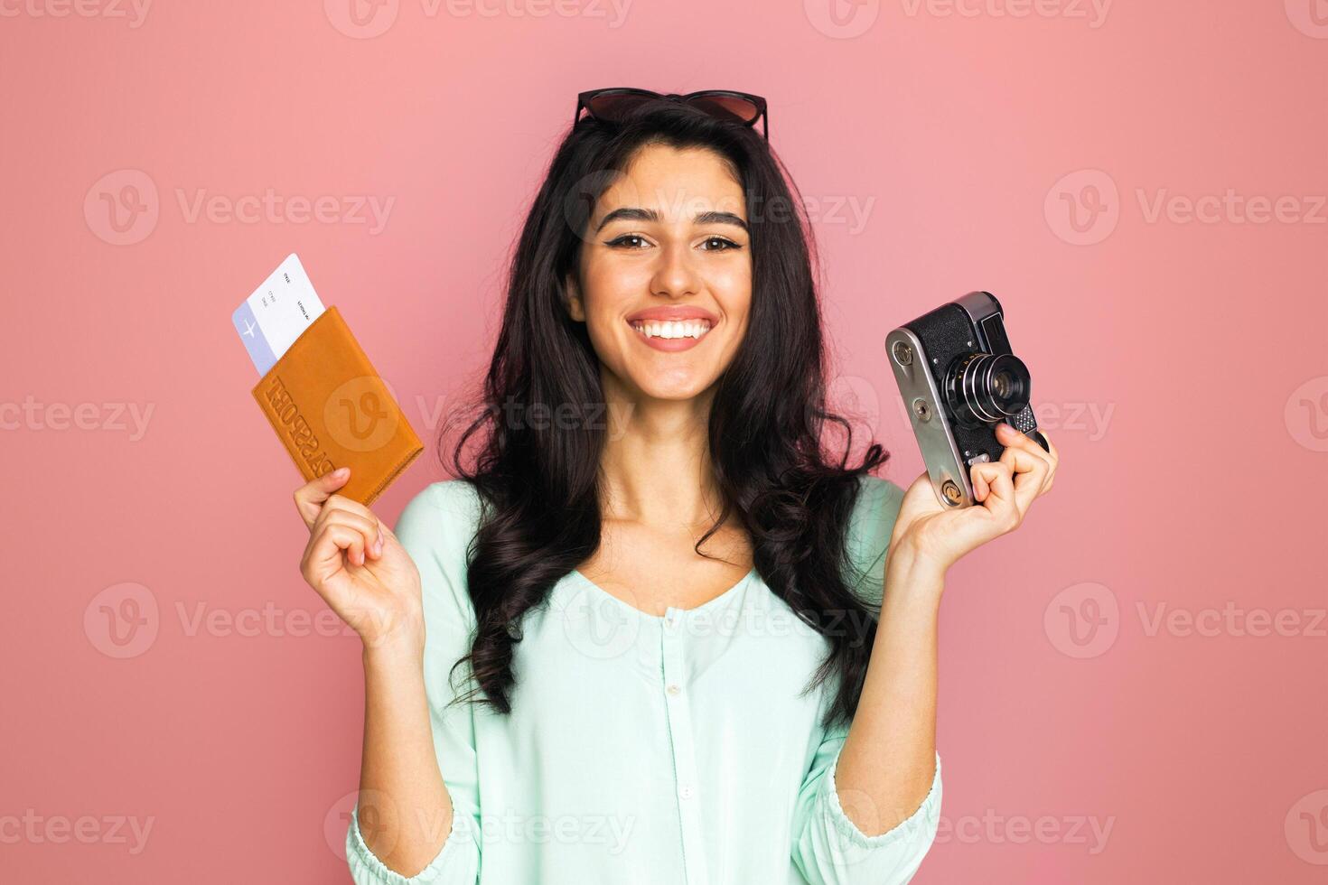 A middle eastern woman is standing while holding a camera and a passport. She appears to be ready for traveling or documenting her journey photo